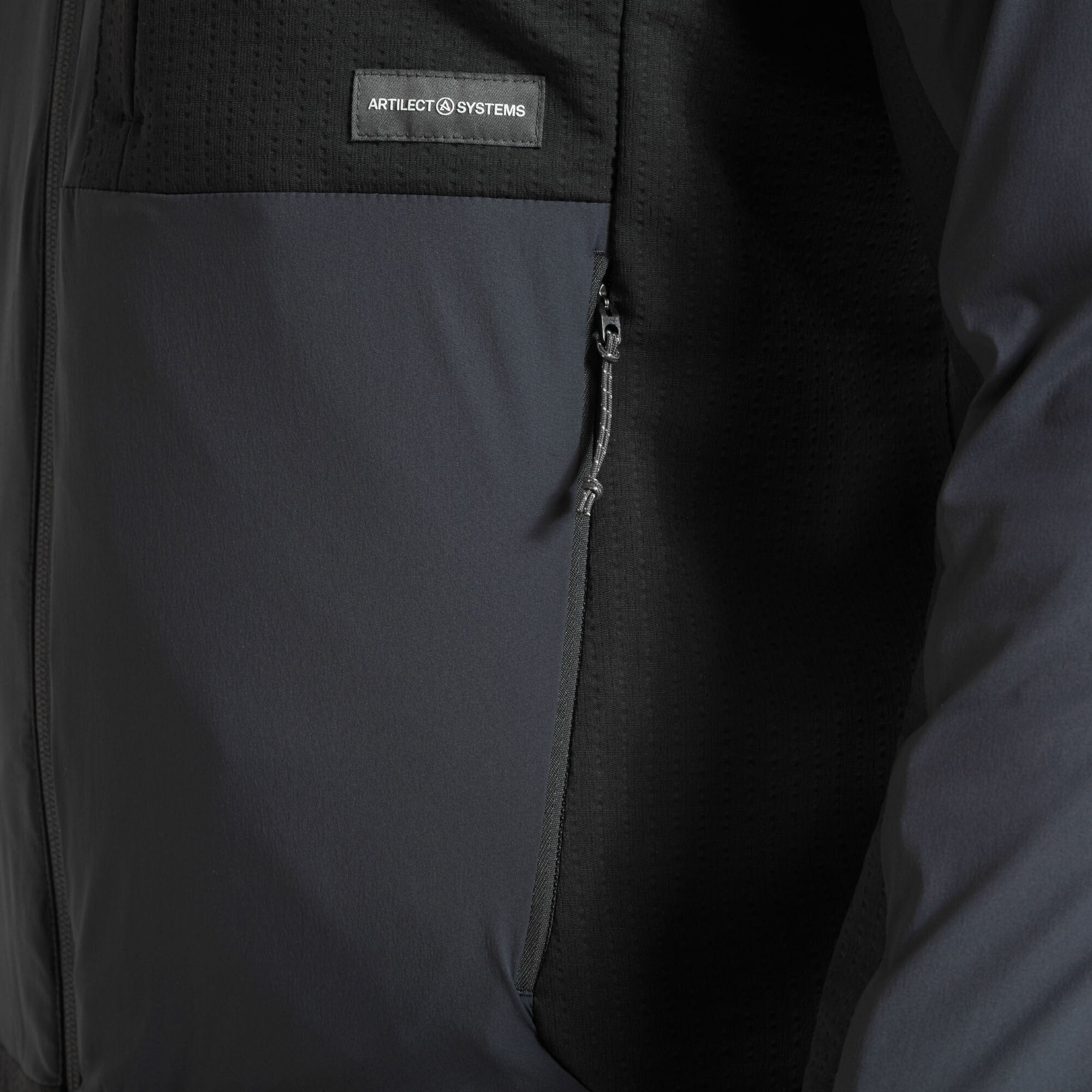 This image zooms in on the chest pocket area on the Artilect Performance Mens Darkstart Fusion Jacket Blue with a clear view of the Artilect Systems logo tag and the durable zipper construction of the hand pocket below.