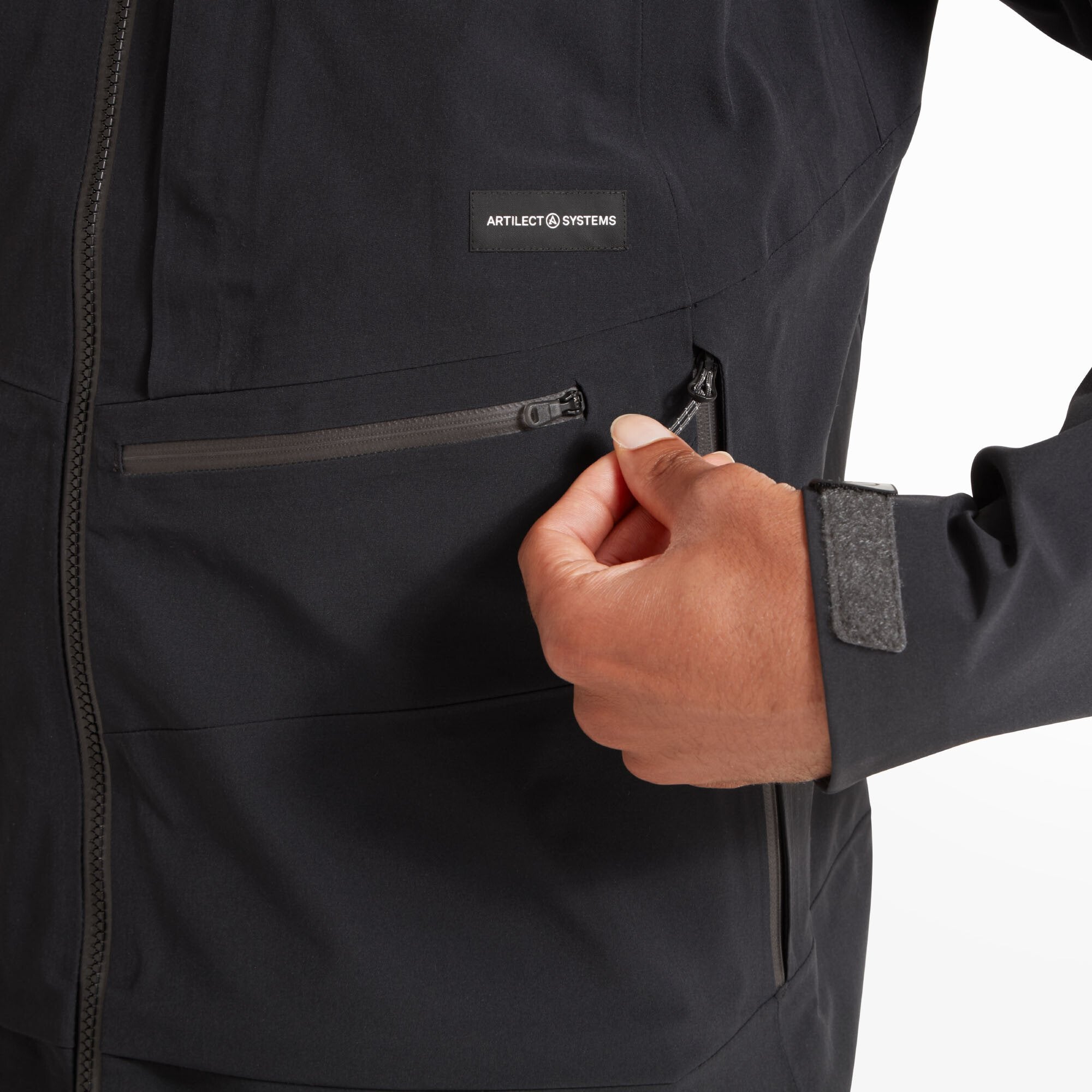 A zoomed-in view of the lower pocket area where the man is unzipping one of the side hand pockets on the Artilect Performance Mens Formation 3L Jacket Black. A logo patch reading “Artilect Systems” is stitched above the pocket.