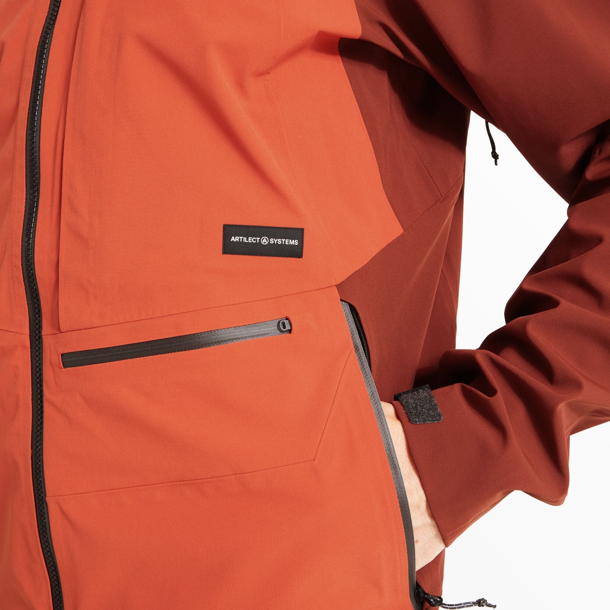 The model's right hand is partially inserted into a zippered lower pocket on the Artilect Performance Mens Formation 3L Jacket Red. The image highlights the matte finish of the fabric, the angled waterproof zipper, and the Artilect Systems logo patch just above it.
