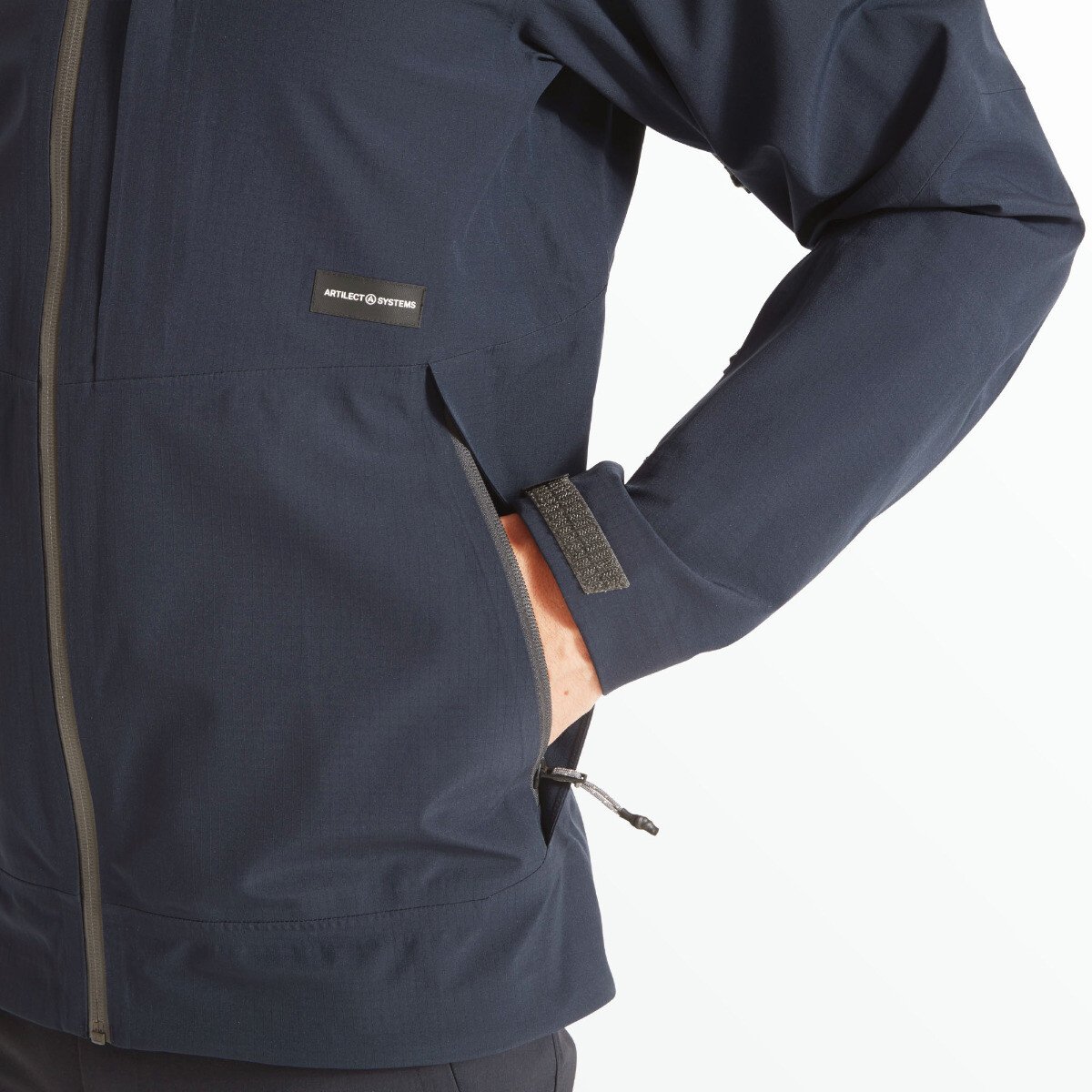 A close-up of the left side of the Artilect Performance Mens Grandview 3L Jacket Blue showing the model's hand inside a zippered front pocket. A small black patch reading "ARTILECT SYSTEMS" is visible above the pocket, and an adjustable Velcro wrist strap is partially fastened.