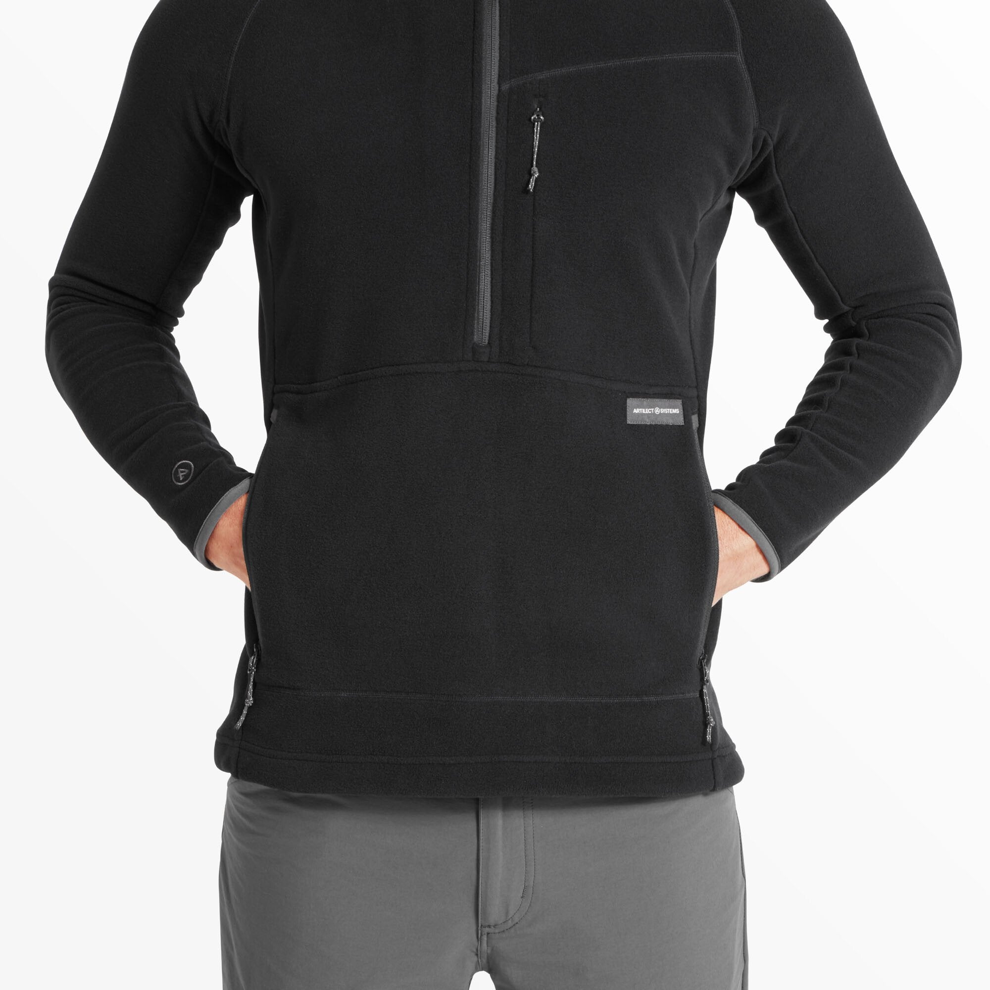 This shot captures the model’s hands partially tucked into the front zippered pockets on the Artilect Performance Mens Halfmoon Bio Pullover Black. The image highlights the “Artilect Systems” brand label on the left pocket and shows the curved seam design around the waist area.