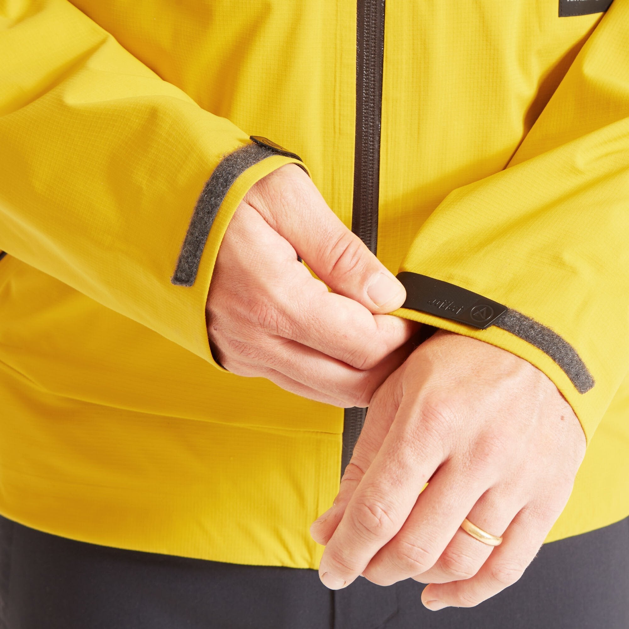 Close-up of the model adjusting the Velcro cuff of the Artilect Performance Mens Shadow Canyon Jacket Yellow sleeve. The Velcro tab features the Artilect logo. This detail showcases the adjustable fit at the wrist.
