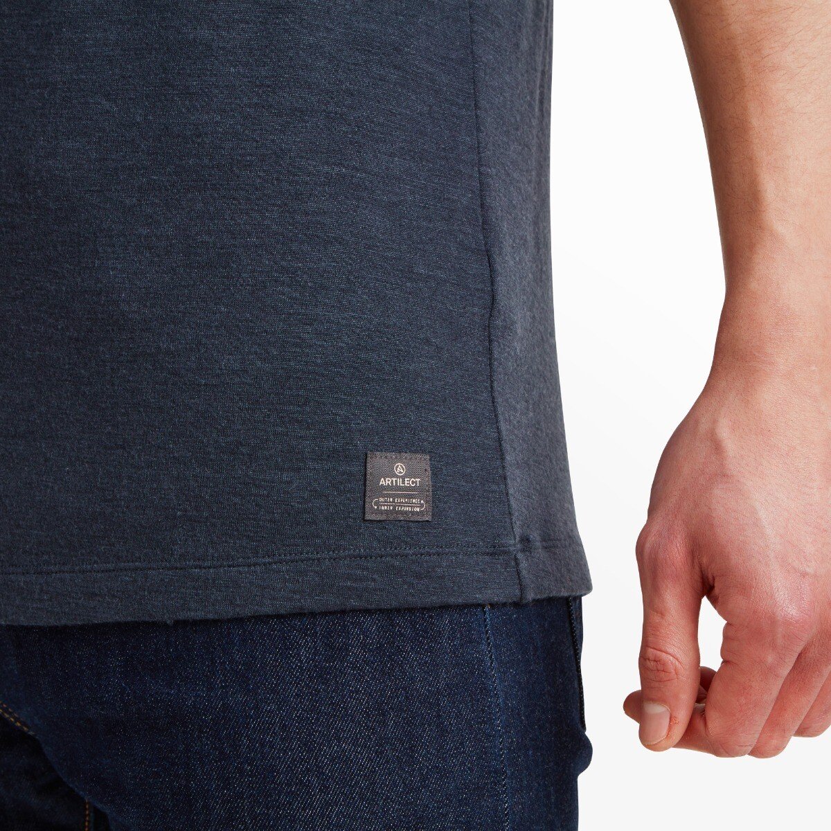 A close-up of the lower right side of the Artilect Performance Mens Utilitee Draft Blue, showing a small black fabric tag sewn onto the hem. The tag displays the Artilect logo and branding in white text. The wearer’s hand and dark denim jeans are partially visible.