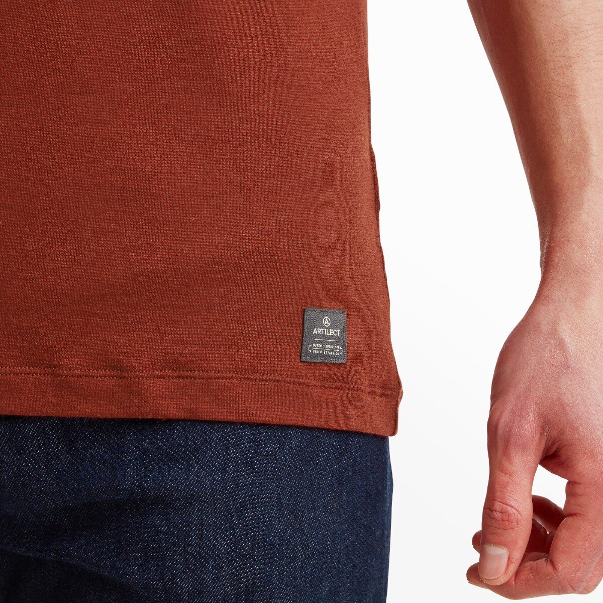 A close-up of the lower left side of the Artilect Performance Mens Utilitee Stacked Orange hem, highlighting a small black woven label that reads “ARTILECT” with extra branding details, adding a technical aesthetic. The model's hand and part of the dark denim waistband are also visible.