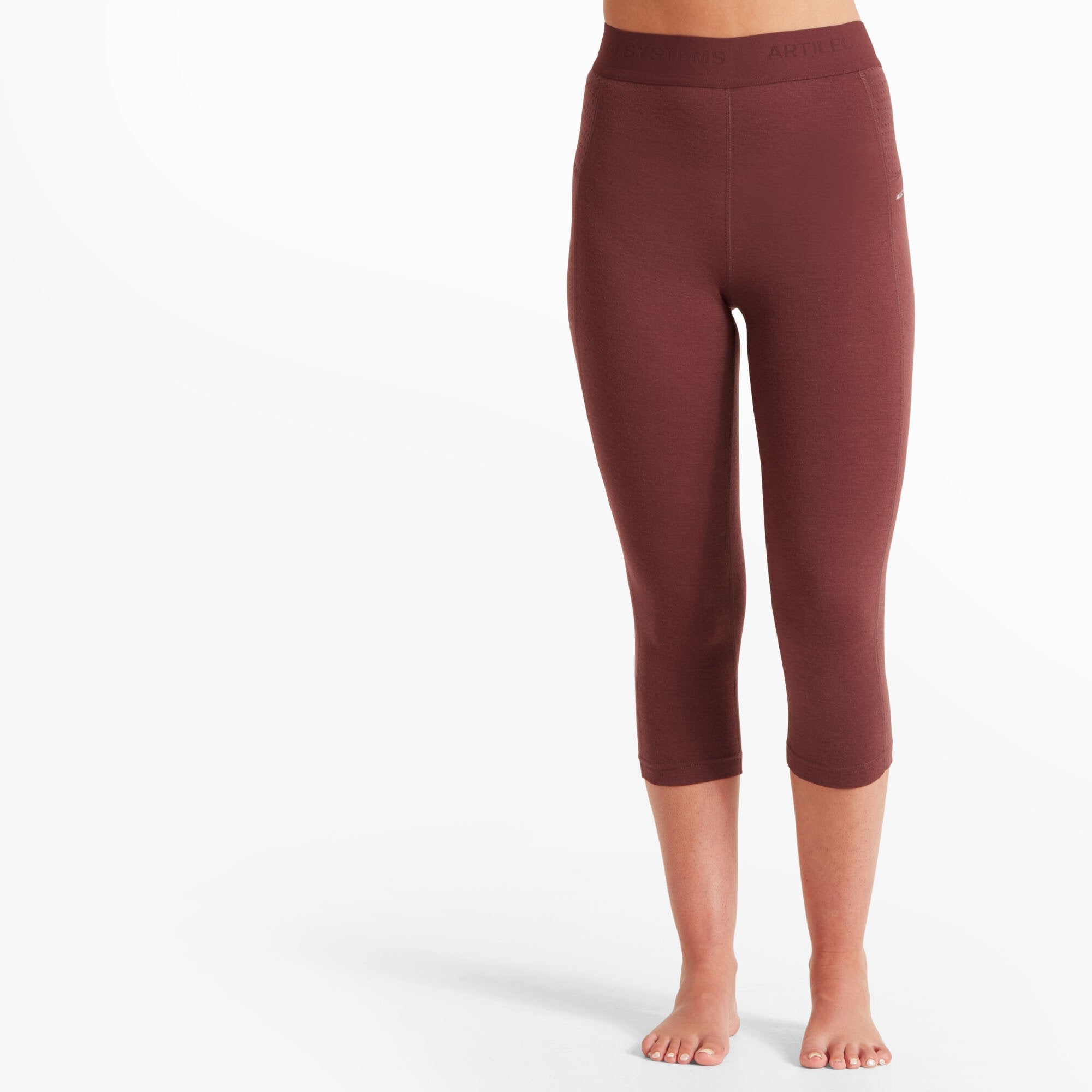 A close-up shot of a woman’s lower body wearing Artilect Performance Womens Boulder 125 3/4 Legging Burgundy. The leggings have a snug, high-waisted fit with a wide elastic waistband. The model is barefoot, and the leggings end just below the knee, revealing her lower calves and feet. The fabric appears soft and stretchy.