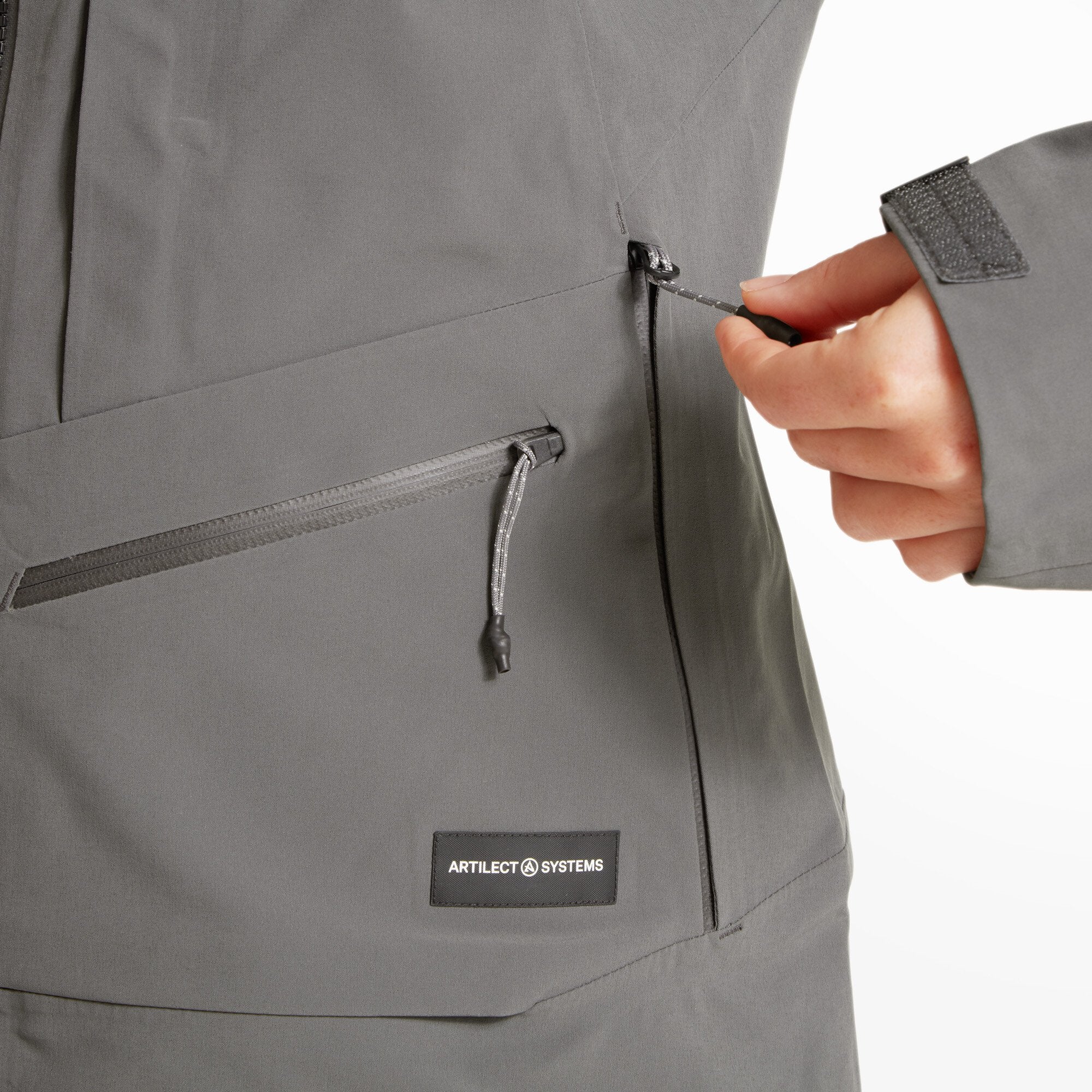 A zoomed-in image of the Artilect Performance Womens Formation 3L Jacket Grey’s right side, focusing on two vertical zippered pockets. A hand is adjusting the bottom zipper pull. The Artilect Systems logo patch is visible above the hem.