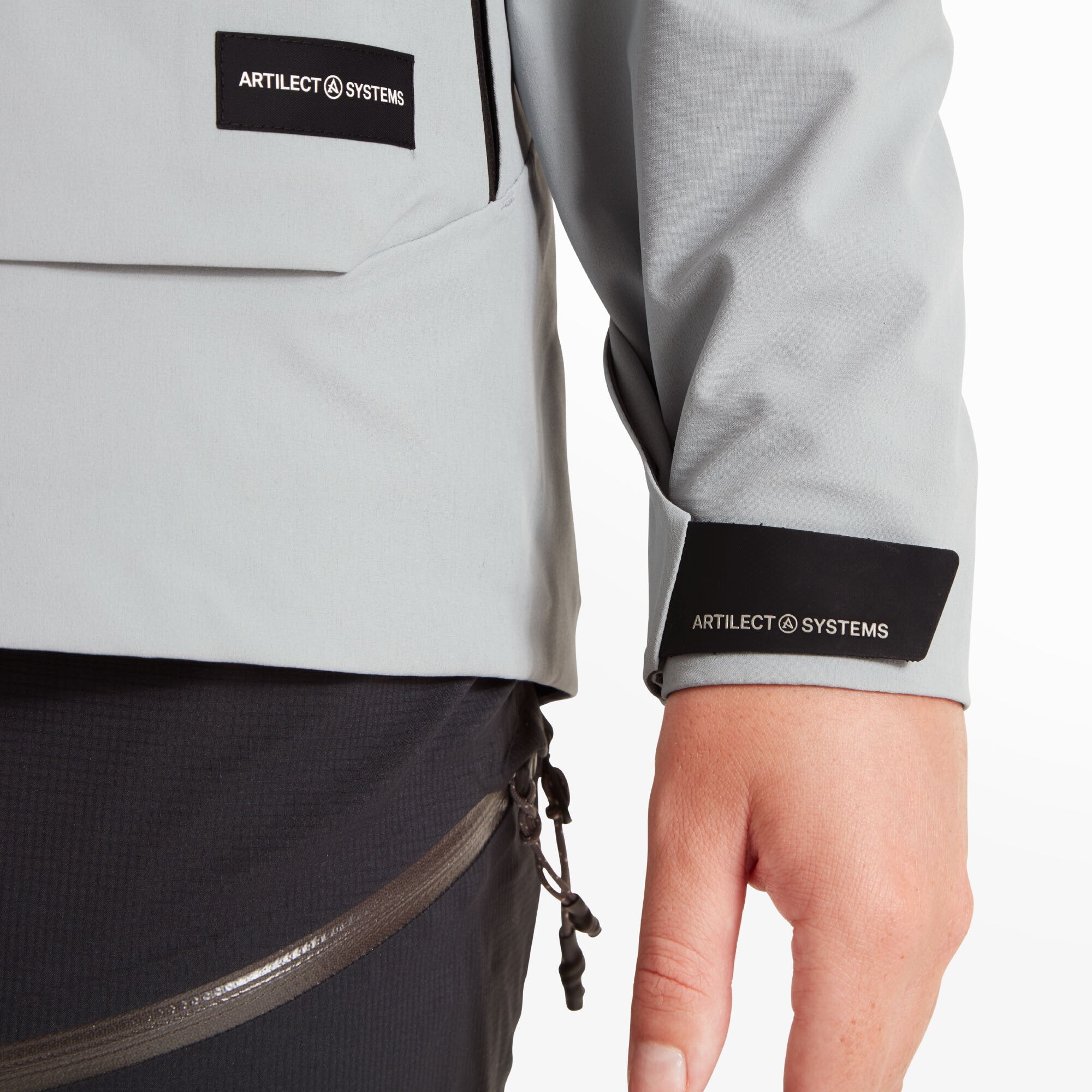 Close-up of the lower part of the Artilect Performance Womens Formation 3L Jacket Grey showing the Artilect Systems logo on the wrist strap and the slanted hand pocket zipper; Velcro wrist adjusters are visible.