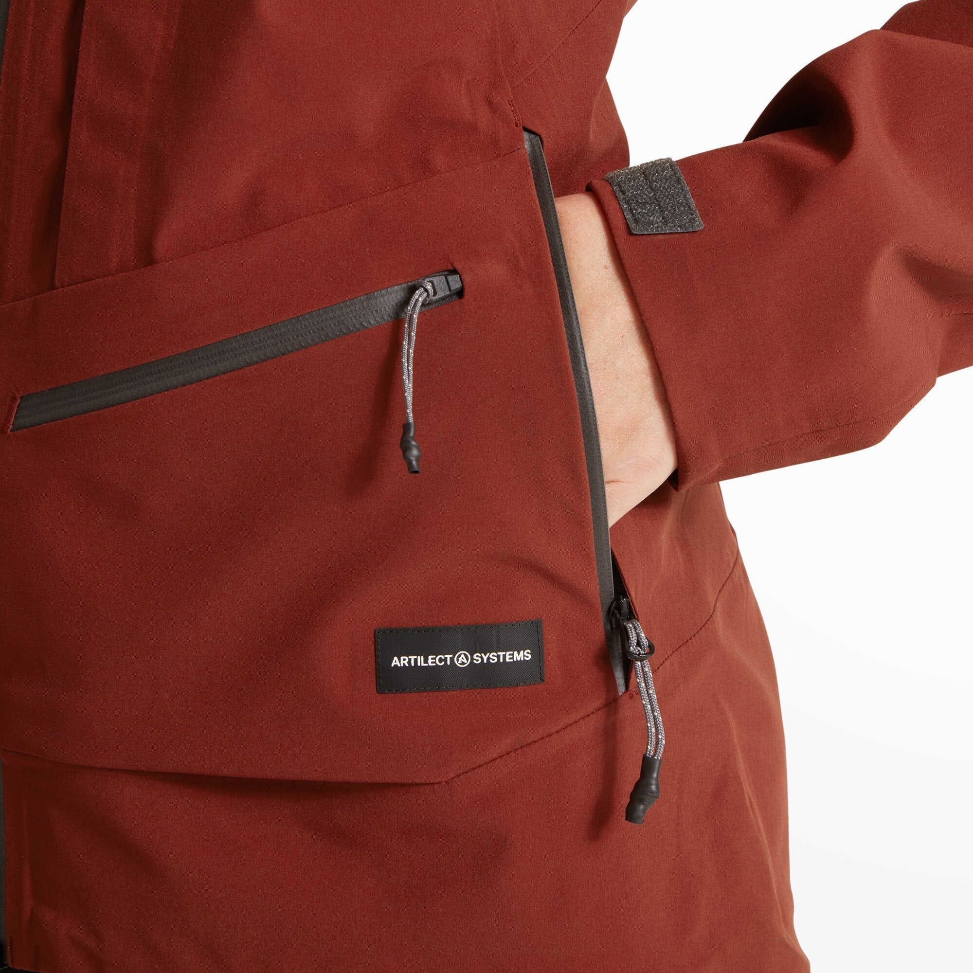 A detailed view of the right hand pocket area of the Artilect Performance Womens Formation 3L Jacket Orange; the model’s hand is inserted into the pocket, and the Artilect Systems brand patch is visible just above the hemline.