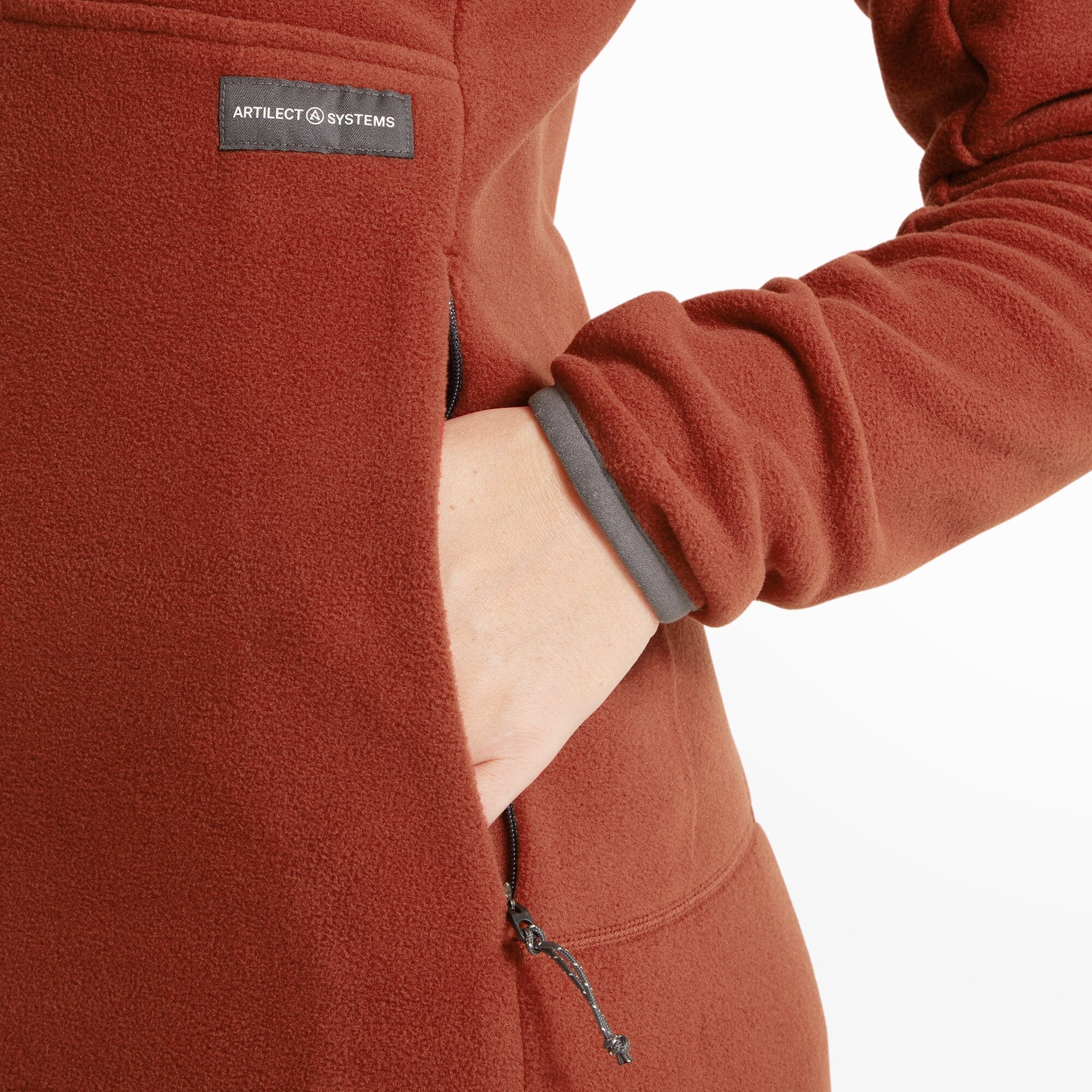 A zoomed-in image of the right side of the Artilect Performance Womens Halfmoon Bio Pullover Orange showing the model’s hand inside the zippered pocket. The ARTILECT SYSTEMS patch is stitched above the pocket. The inner fleece lining is visible at the cuff, which is rolled back slightly.