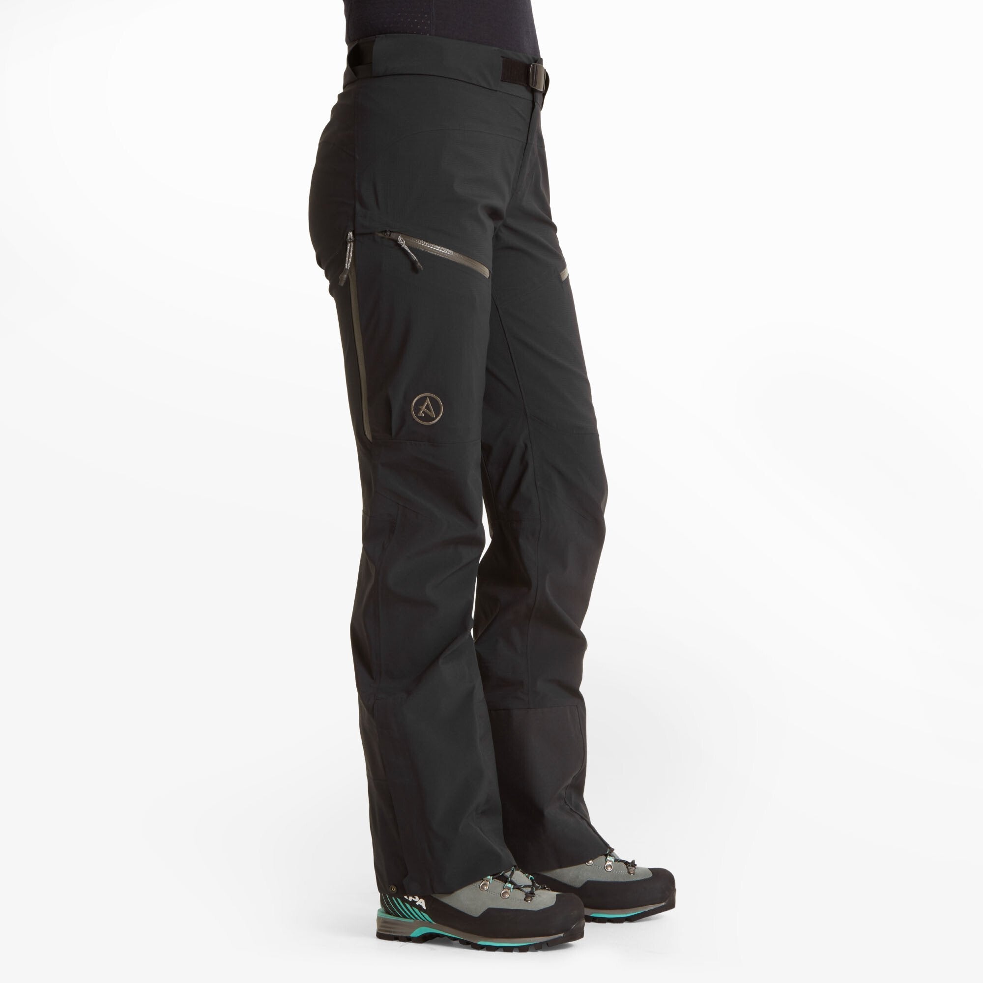 A three-quarter side view of a woman wearing the Artilect Performance Womens Kinetic Pant Black. The image showcases the pant's contoured fit, articulated knees, and dual zippered thigh pockets. The pants feature the Artilect logo on the left thigh and reinforced material near the ankle. The model is wearing grey and teal hiking boots.