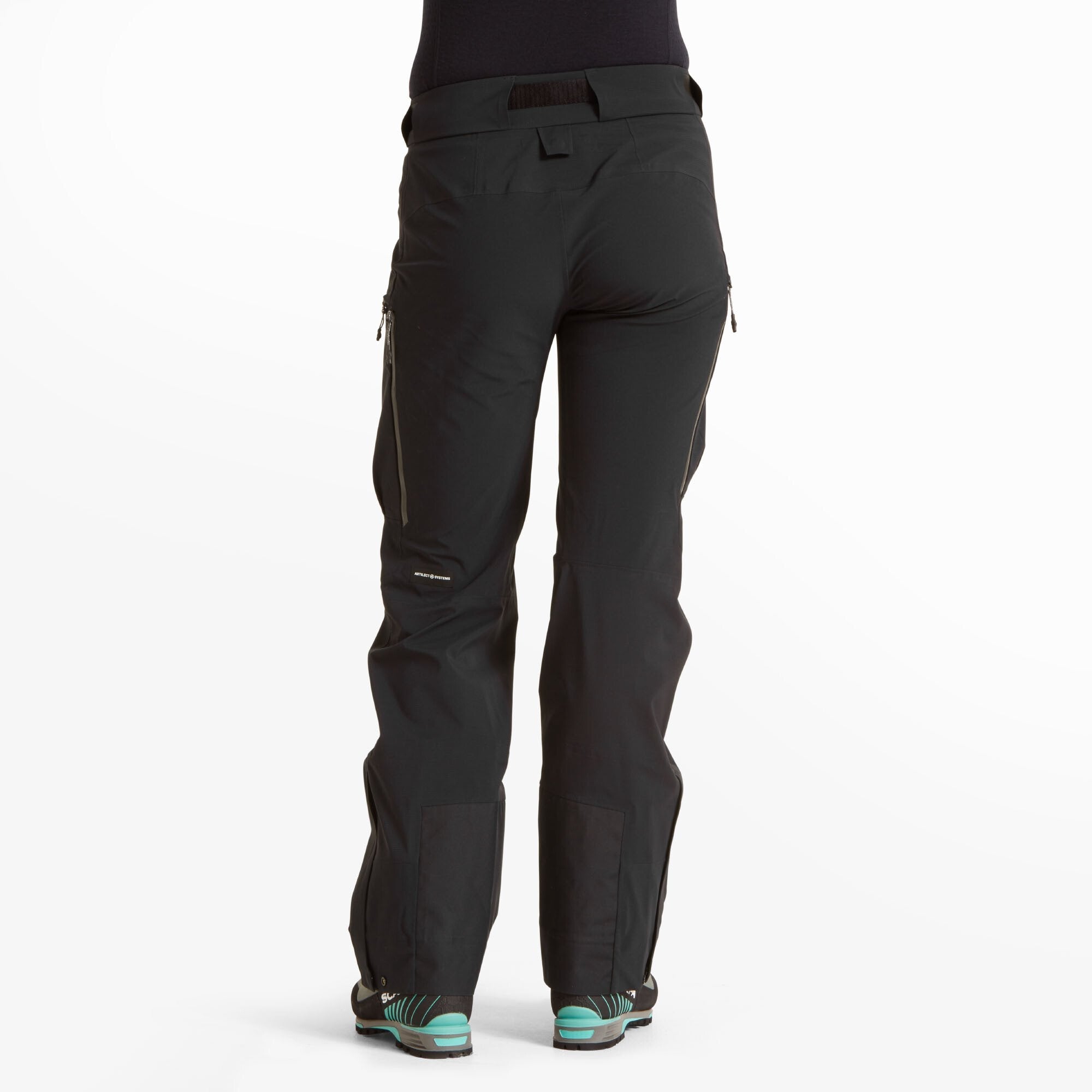 A rear view of the Artilect Performance Womens Kinetic Pant Black, worn by a woman standing with feet shoulder-width apart. The pants feature a secure, mid-rise waistband with belt loops and a wide velcro strap for adjustment. Subtle seam detailing enhances fit and mobility, while zippered rear thigh vents add ventilation.
