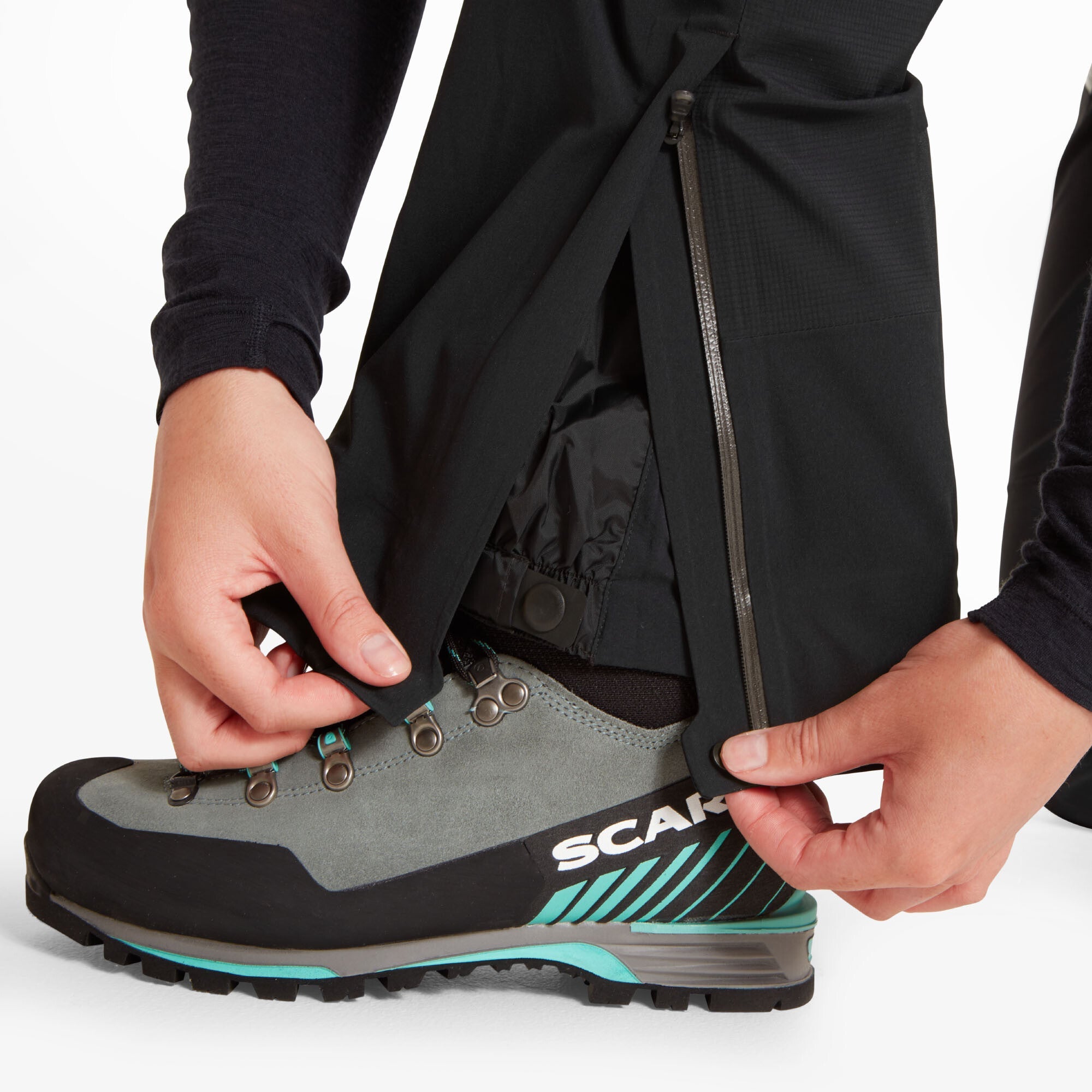 A detailed view of the lower leg and ankle of the Artilect Performance Womens Kinetic Pant Black as a person adjusts the built-in internal gaiter over their hiking boot. The image reveals a vertical zipper at the ankle for expanding the pant opening, and an internal snow gaiter designed to fit snugly over boots, ideal for alpine activities.