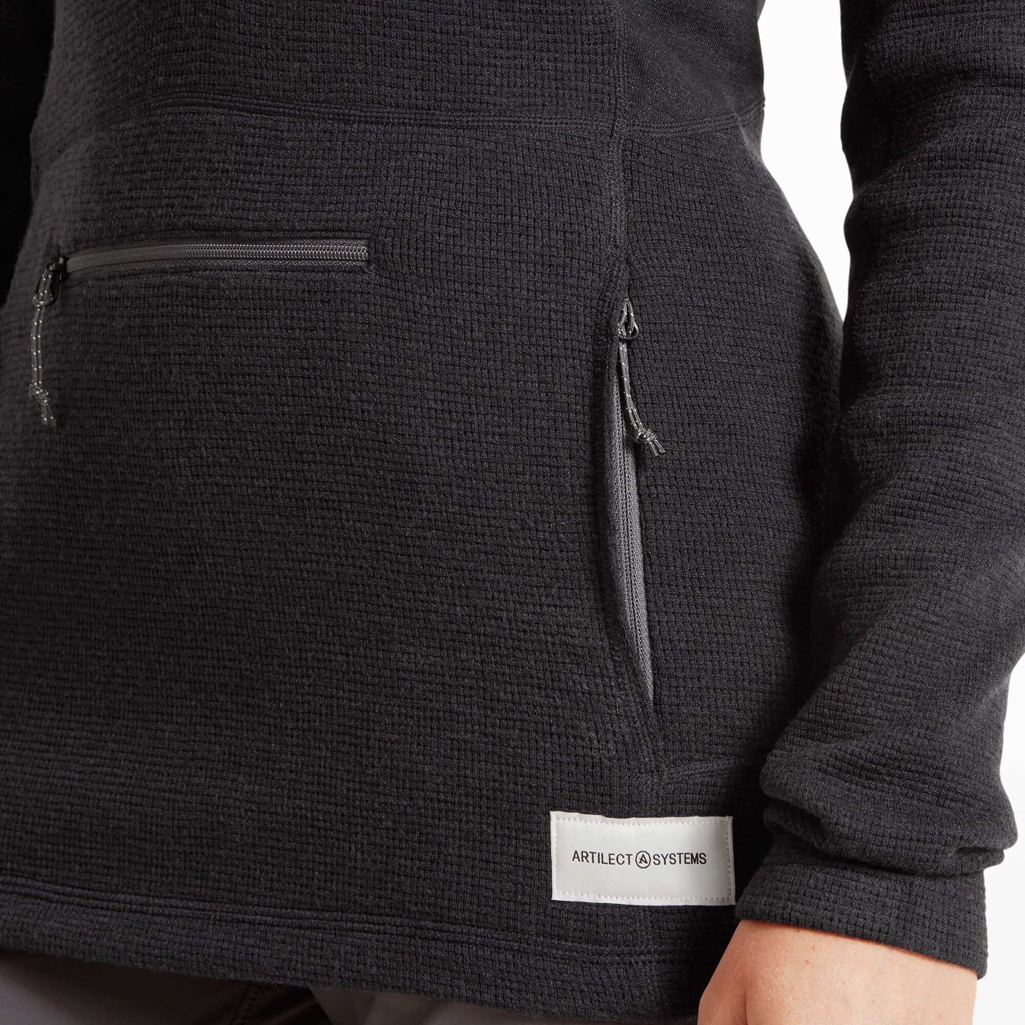 Close-up of the Artilect Performance Womens Sundown 250 Half Zip Top Black’s lower front section showing two zippered hand pockets and a small white ARTILECT SYSTEMS logo label sewn near the hem.