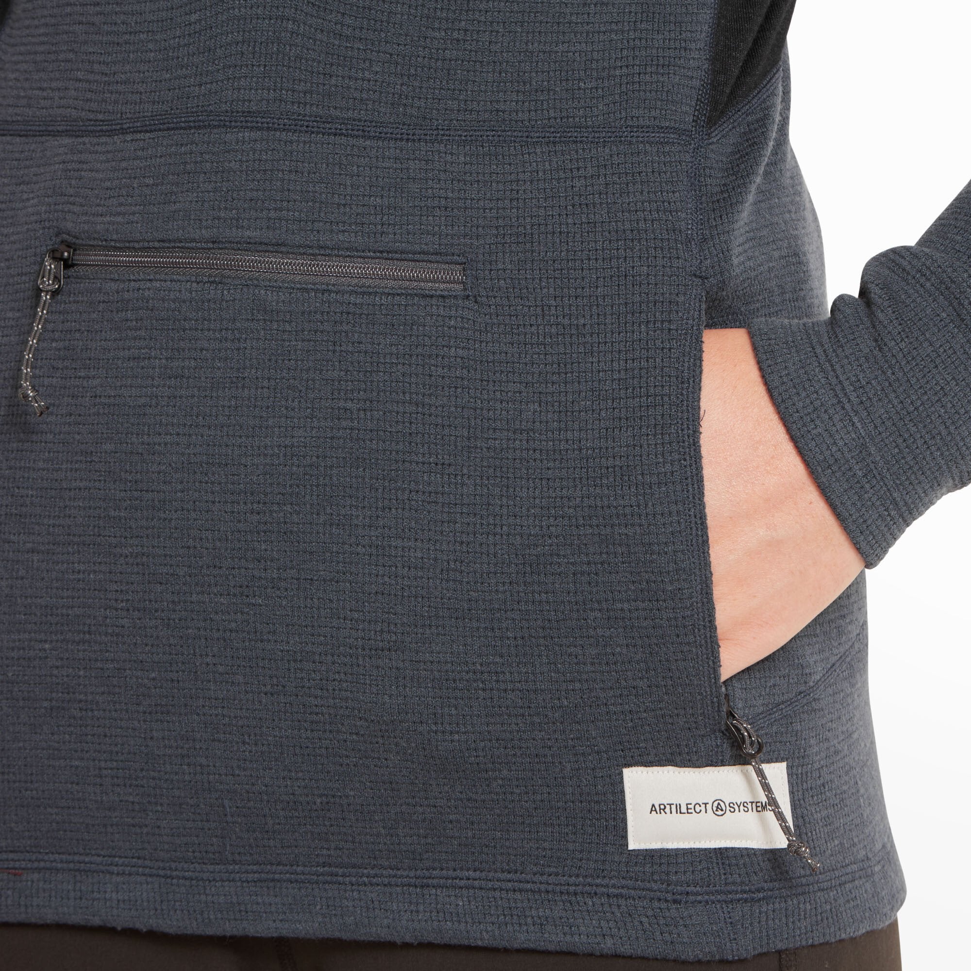A close-up view of the kangaroo pocket at the front of the Artilect Performance Womens Sundown 250 Half Zip Top Blue. One hand is slipped inside a side-entry pocket while the other zipper pull is visible, showcasing the utility of the double-entry pocket system. The Artilect Systems logo is printed on a white label at the lower right hem.