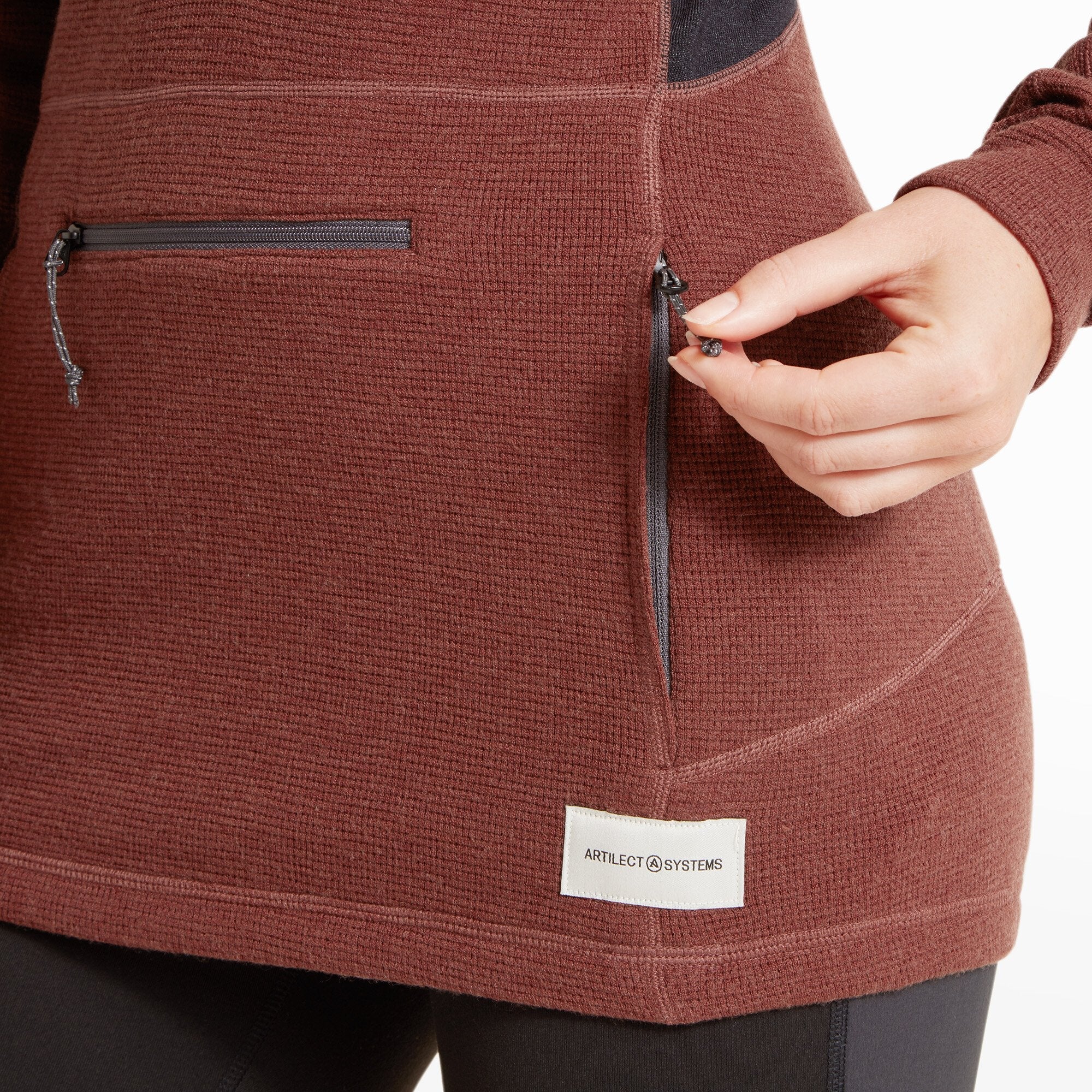 A detailed close-up of the model’s hands, showing the extended sleeves with thumbholes on the Artilect Performance Womens Sundown 250 Half Zip Top Burgundy. The burgundy textured fabric wraps around the hand, with a black interior fabric barely visible underneath. The fit of the cuff is snug, offering a secure feel for active wear.