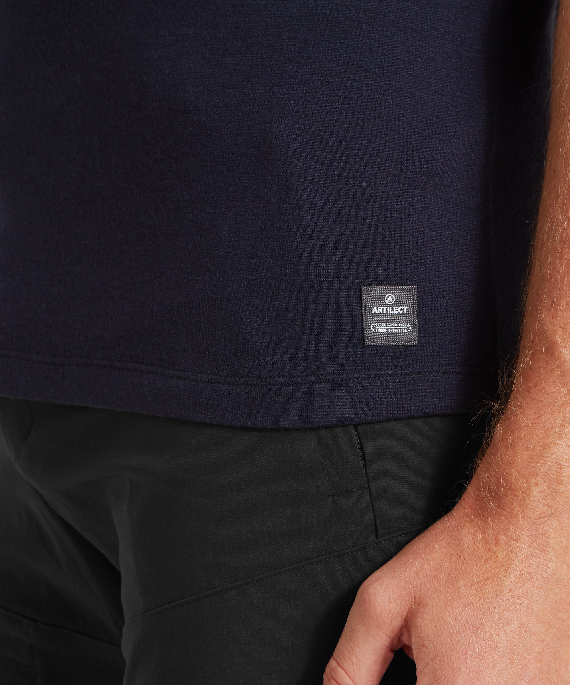 A close-up of the lower hem of a navy T-shirt featuring a small rectangular ARTILECT brand label, paired with black trousers.