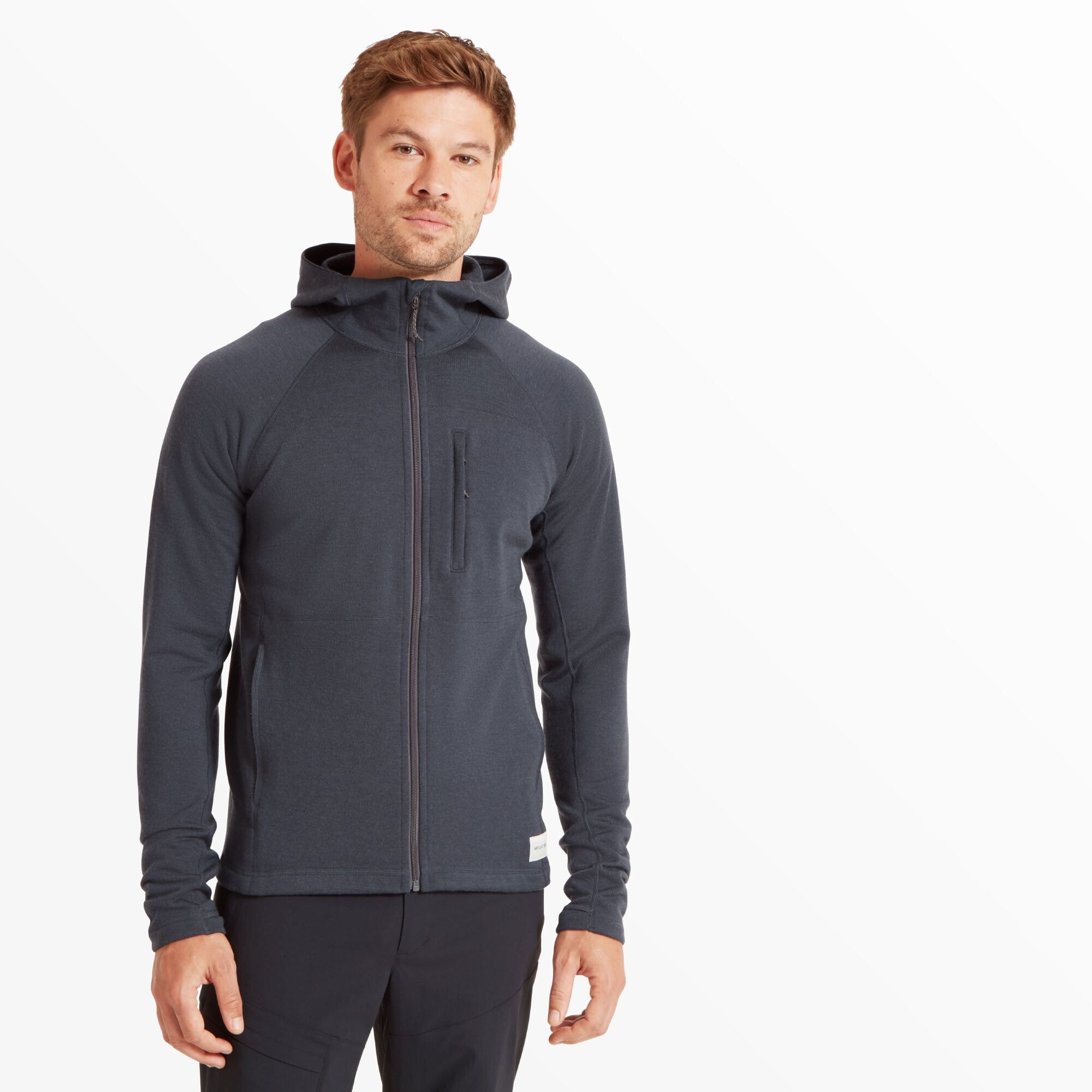 A man is wearing the Artilect Performance Mens Eldorado Merino Hoodie Blue. The hoodie has a full front zipper, a slim fit, and features a zippered chest pocket on the left side. The hood is down, and the model is looking slightly to the side with a neutral expression.