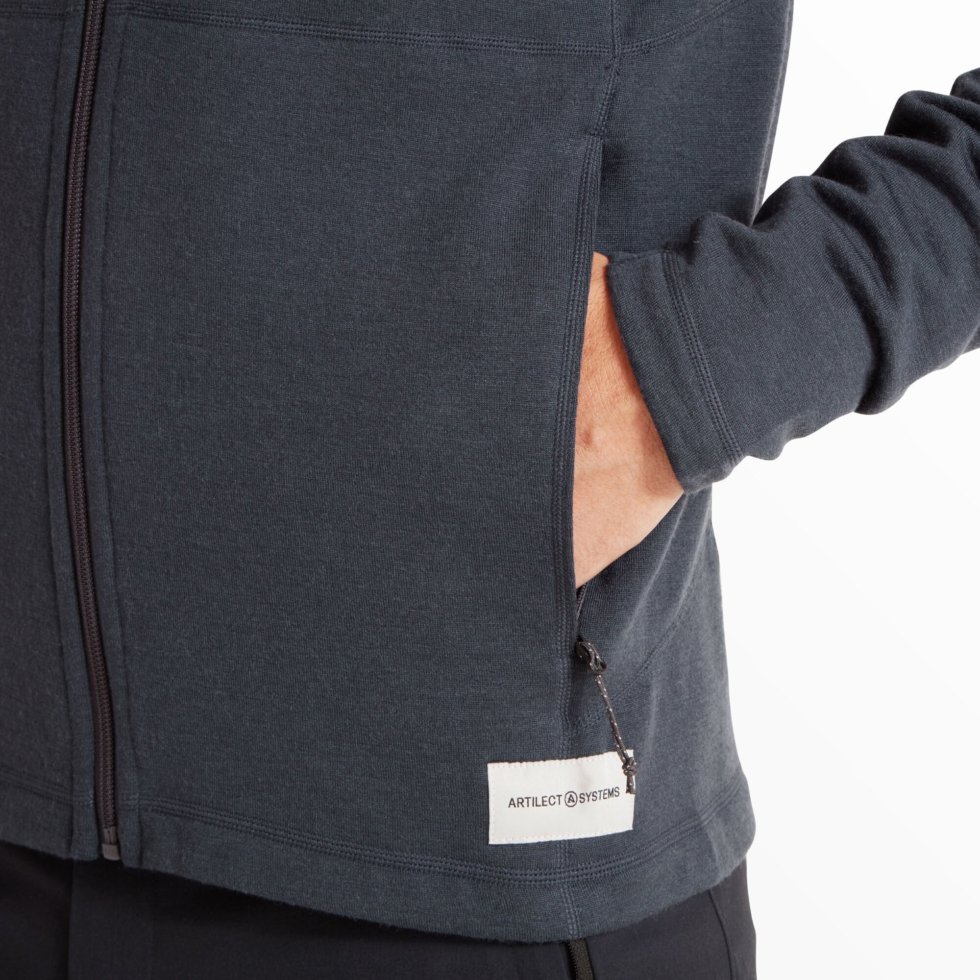 The lower portion of the Artilect Performance Mens Eldorado Merino Hoodie Blue is shown with the model’s hand partially inserted into one of the side pockets. The image reveals a small white woven label near the hem with the text "ARTILECT SYSTEMS" printed on it, along with subtle seam detailing around the pocket.