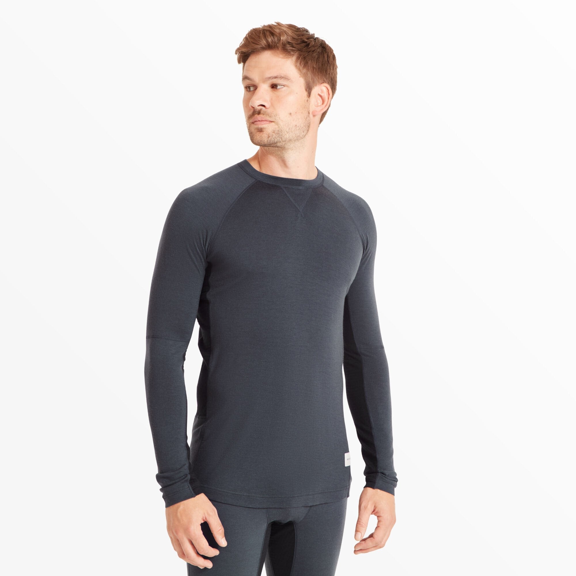 A male model is standing facing forward wearing a fitted, long-sleeve, Artilect Performance Mens Flatiron 185 Crew Blue. The shirt features raglan sleeves and a clean, minimal look with no front graphics, ideal for layering in cold-weather activities.