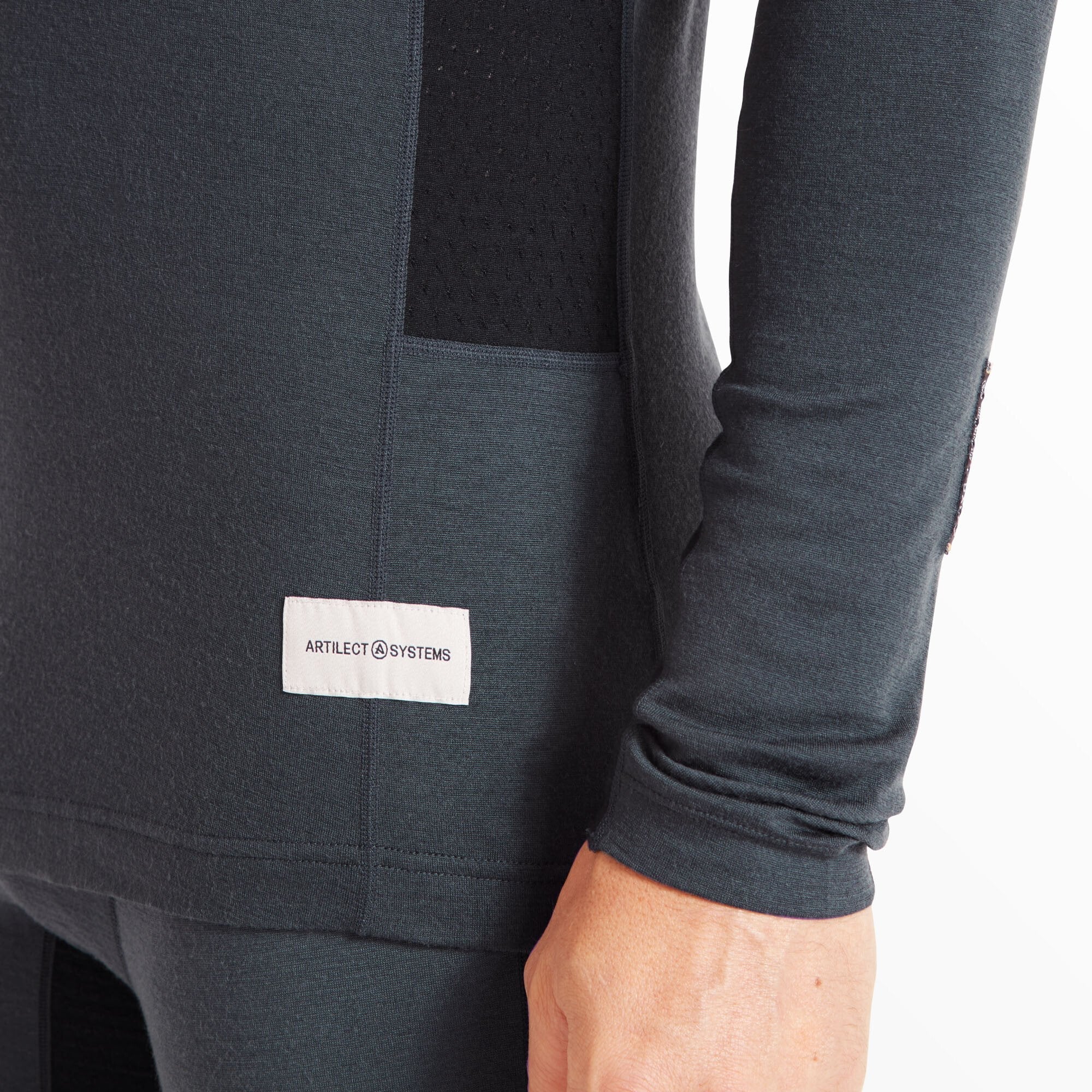 This close-up focuses on the lower side hem of the Artilect Performance Mens Flatiron 185 Crew Blue. A white woven Artilect Systems logo patch is stitched onto the shirt, and breathable mesh side panels are partially visible.