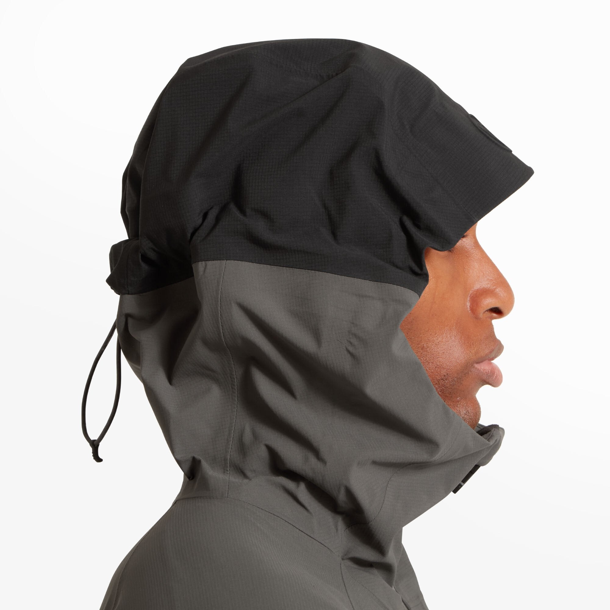 The model is turned to show the back of the Artilect Performance Mens Shadow Canyon Jacket Grey. The hood features a black panel contrasting the rest of the jacket’s dark grey color. The back is plain with minimal seam lines for a clean design.
