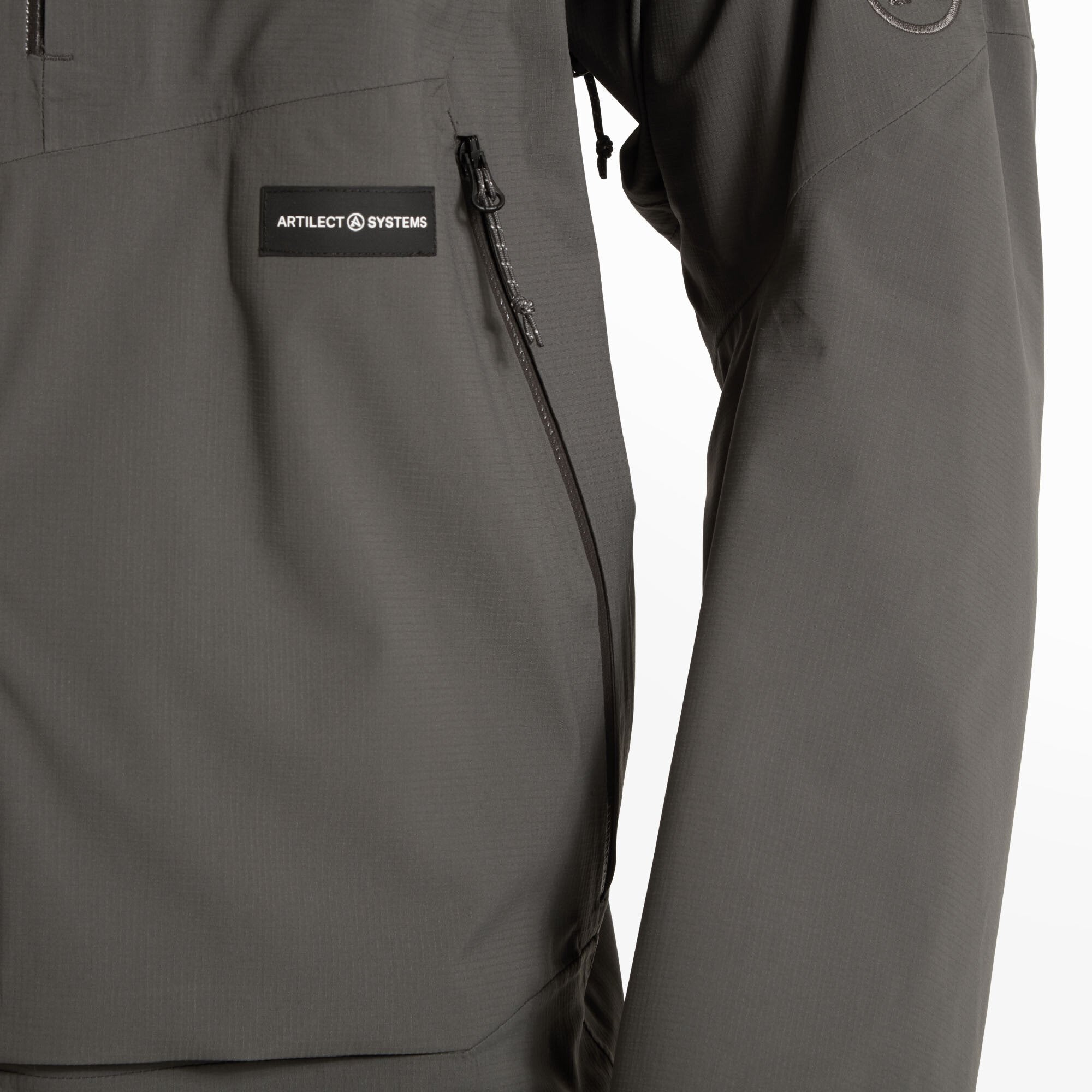 Focused view of the left chest area. The Artilect Systems patch is stitched above a vertical zippered chest pocket. The fabric texture is subtly patterned with a technical look on the Artilect Performance Mens Shadow Canyon Jacket Grey.