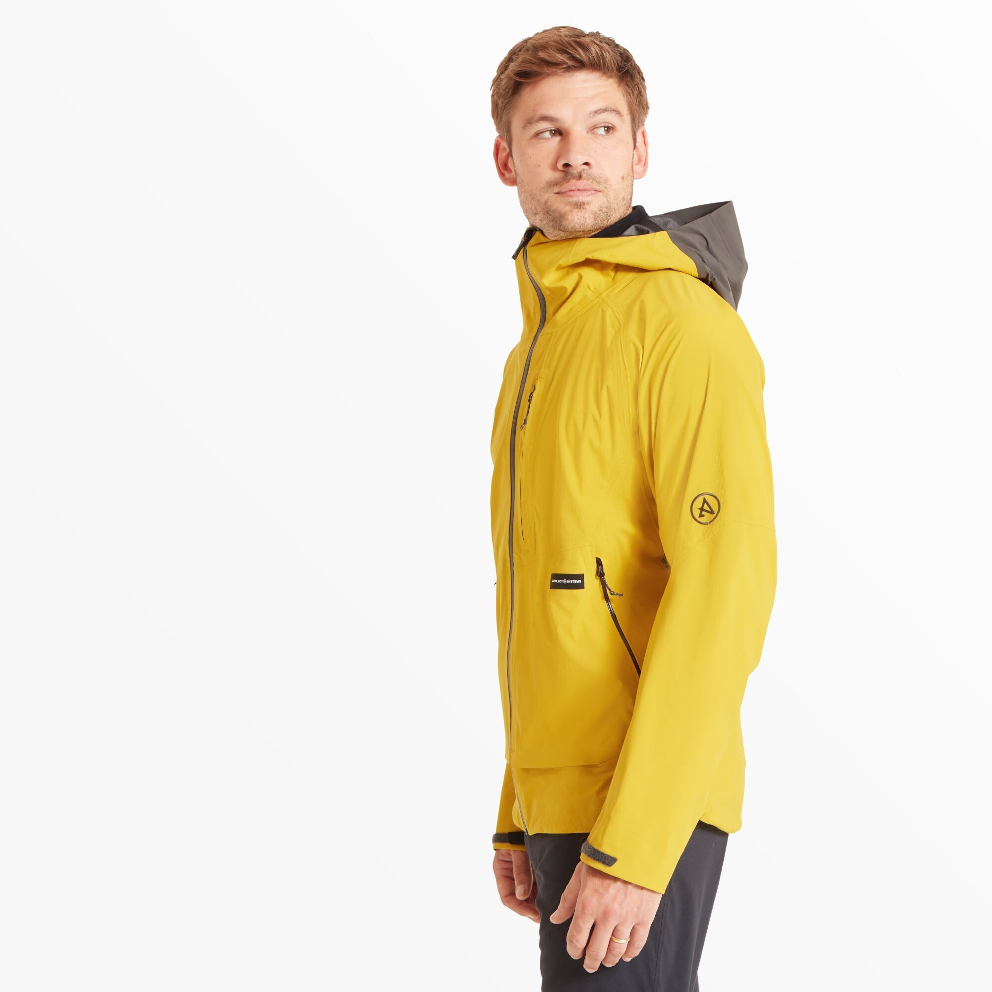 The model is shown from a front-left angle, with his head turned slightly to the left. This view highlights the Artilect Performance Mens Shadow Canyon Jacket Yellow’s side profile, showing the articulated sleeve design and hood construction. The logo patch on the sleeve is clearly visible.