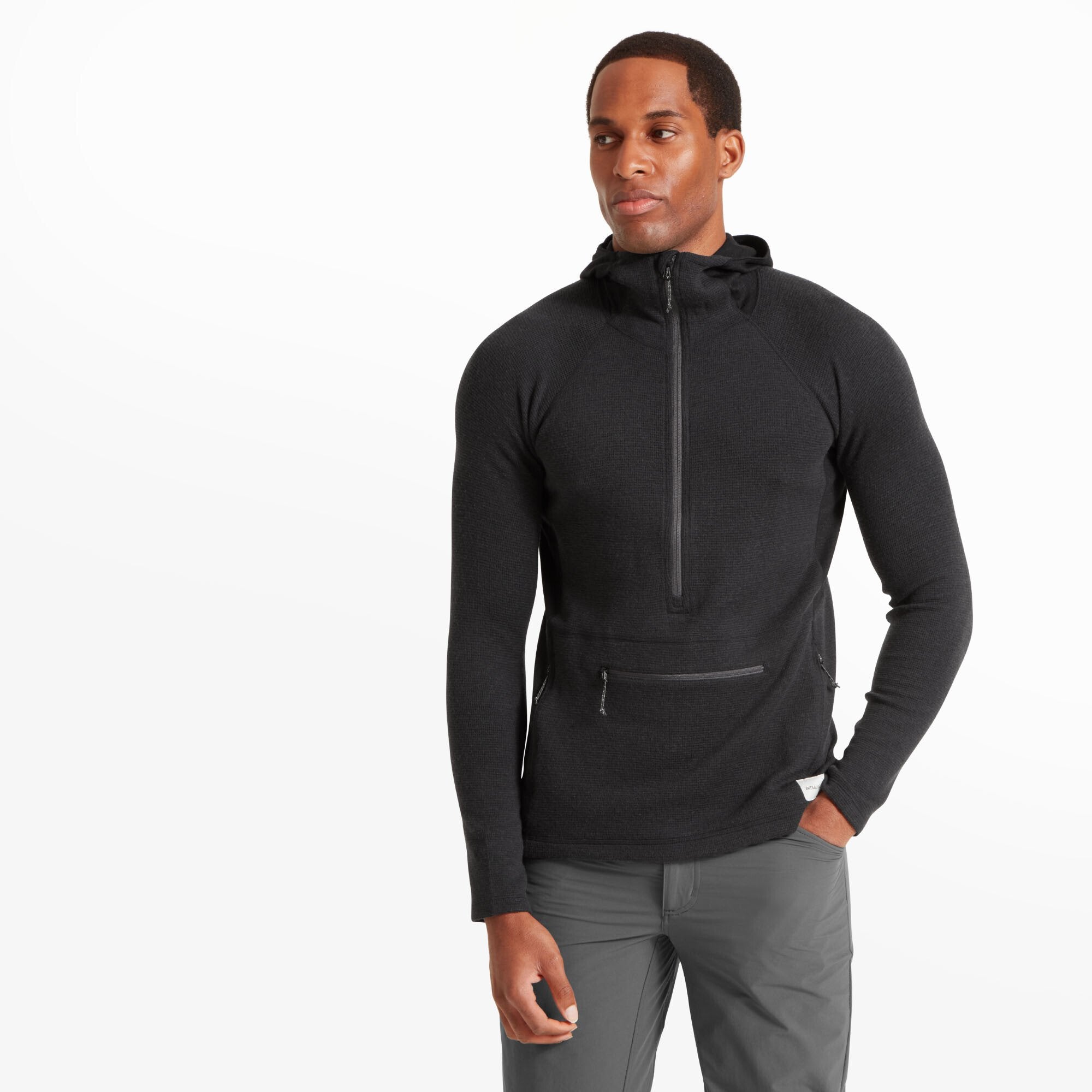 A man stands facing forward, wearing a fitted Artilect Performance Mens Sundown 250 Half Zip Top Black with a hood. The hoodie features a visible half-length zipper and a zippered kangaroo pocket. The texture of the fabric is subtly patterned, and the man has one hand in his pocket, looking slightly to the side.