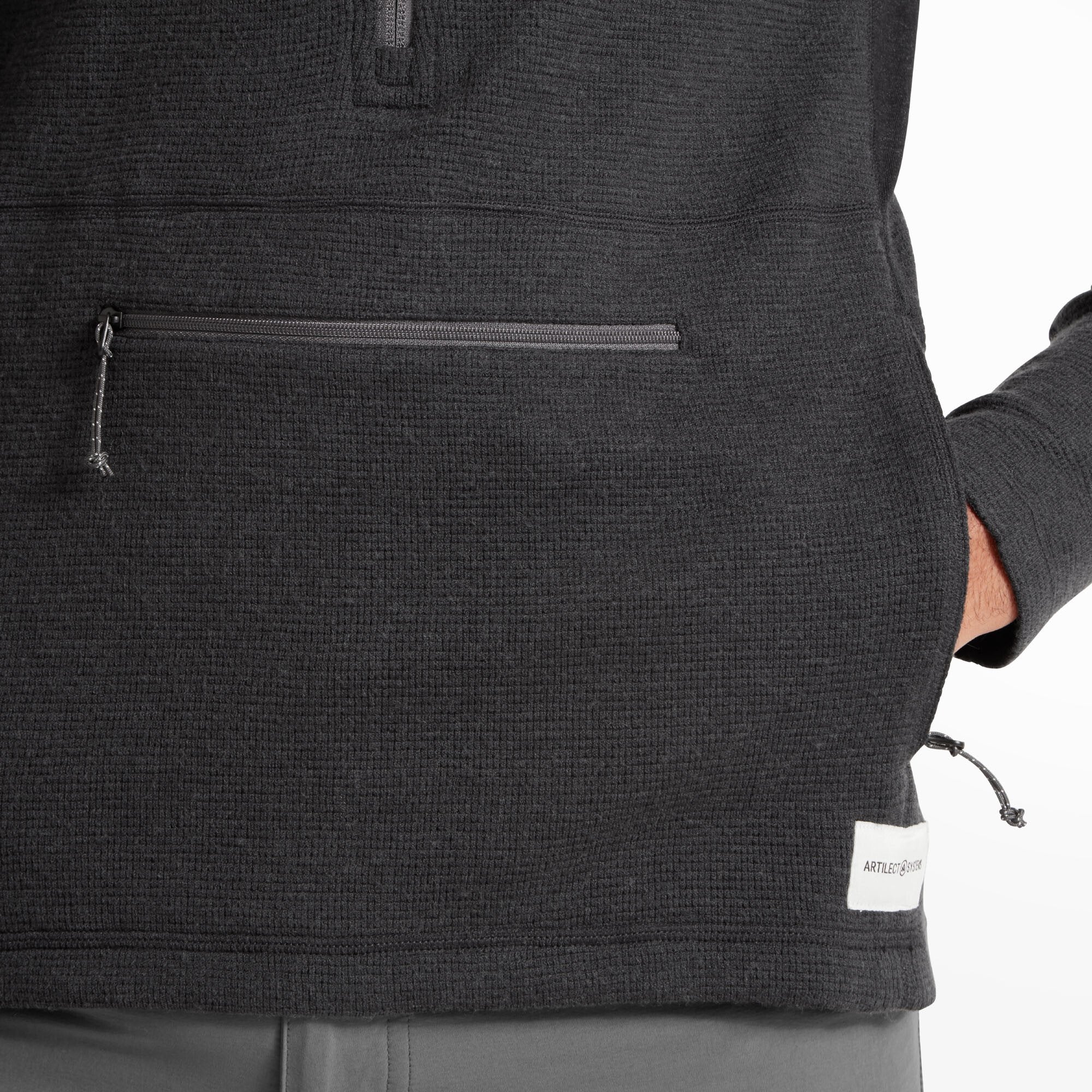 Close-up of the Artilect Performance Mens Sundown 250 Half Zip Top Black’s kangaroo pocket with a horizontal zipper. The model’s hand rests casually in one side. A small “Artilect Systems” label is stitched near the hem.