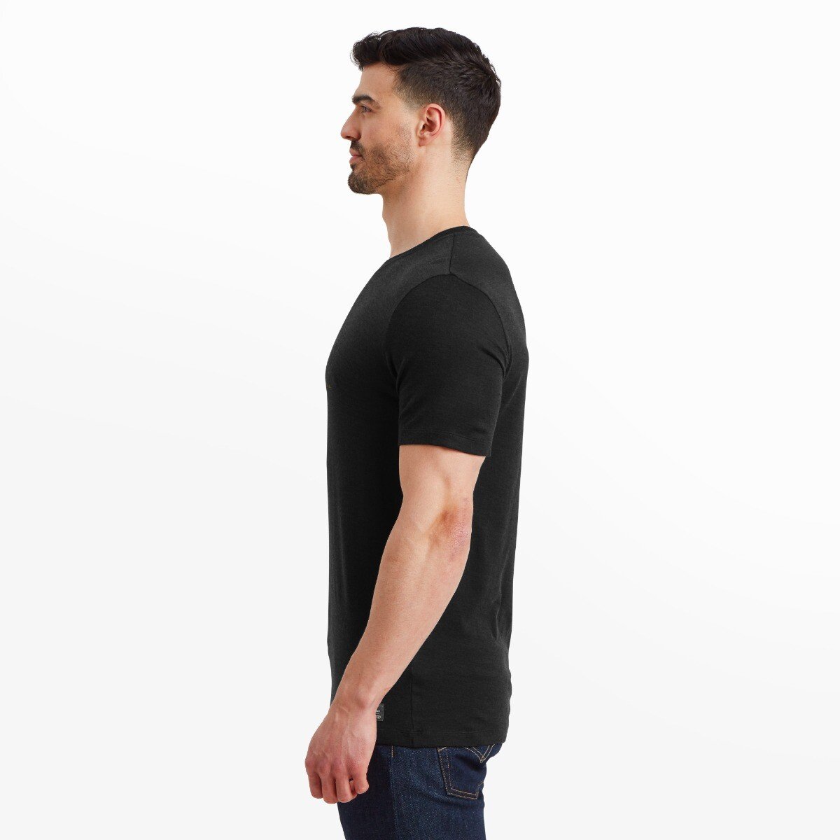 Profile shot of the man wearing the same Artilect Performance Mens Utilitee Draft Black and jeans, standing straight and looking forward. The sleeve and shoulder seam structure are clearly visible, emphasizing the tailored fit of the t-shirt.