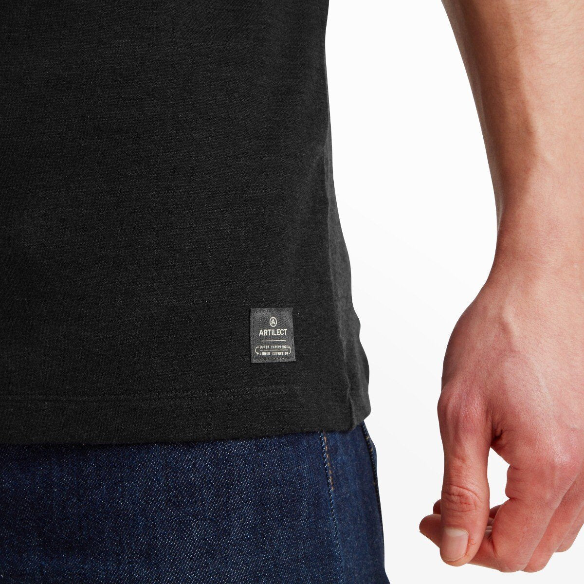 A zoomed-in image of the bottom left hem of the Artilect Performance Mens Utilitee Draft Black, showing a small woven label that reads "Artilect" with branding details. The label is stitched above the seam where the shirt meets the top of the jeans.