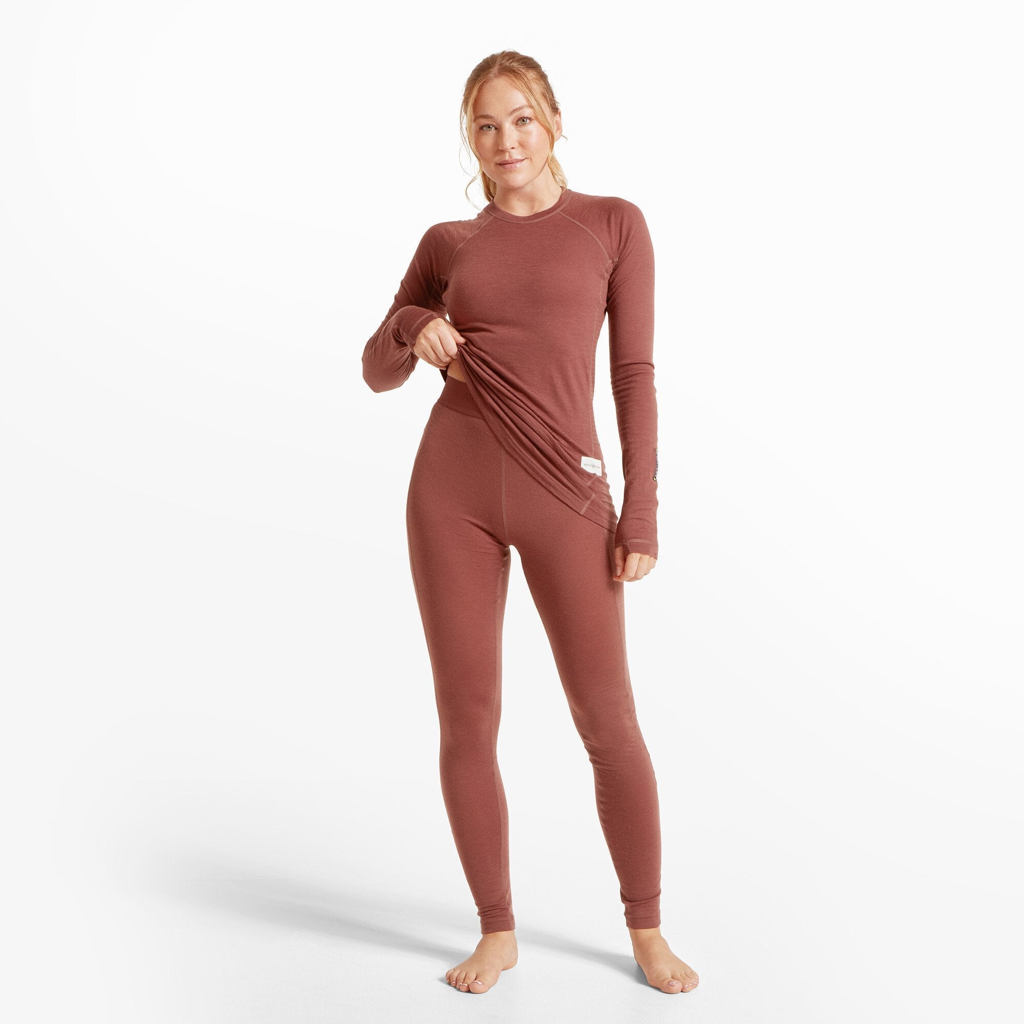 A full-body shot of a woman wearing a matching burgundy-colored long-sleeve top and Artilect Performance Womens Boulder 125 Legging Burgundy. She is slightly lifting the bottom of her top to reveal the waistband of the leggings. She stands barefoot with a relaxed and confident posture.