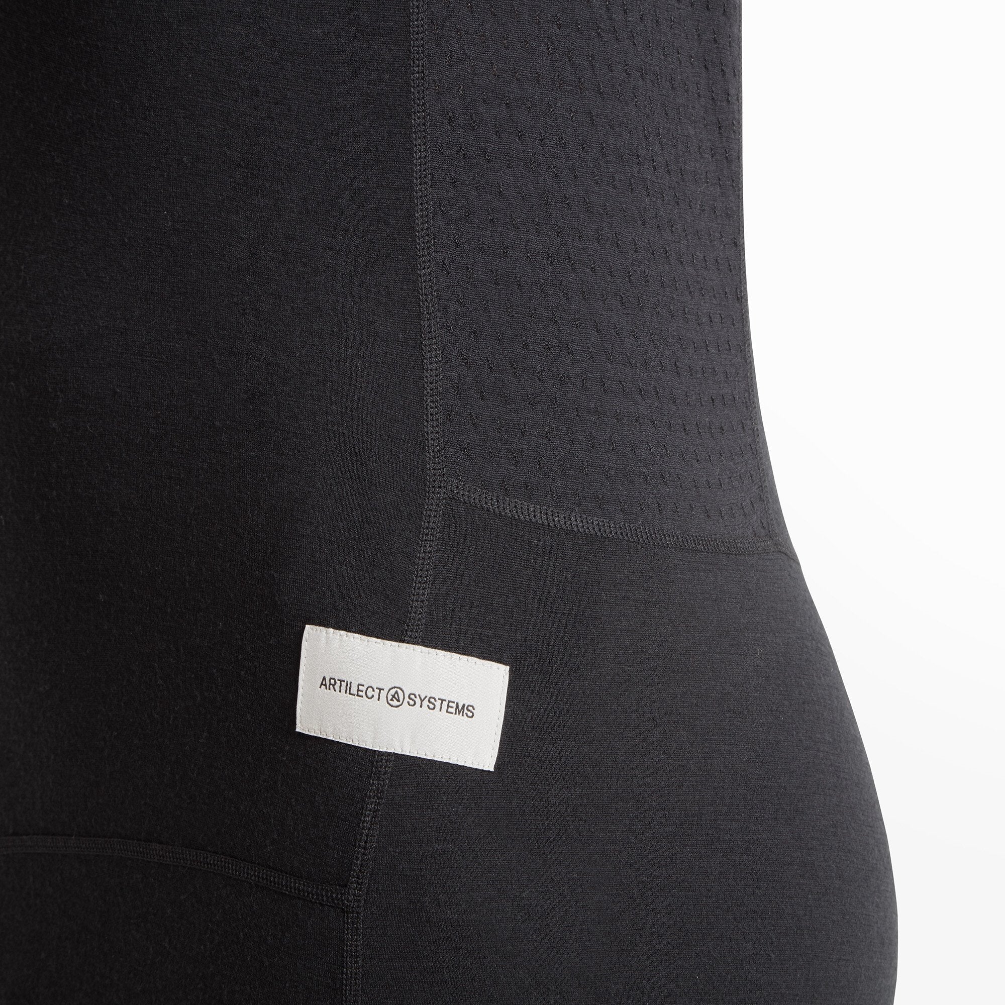 A close-up of the lower side of the Artilect Performance Womens Boulder 125 Quarter Zip Top Black, showing a white rectangular "ARTILECT SYSTEMS" logo patch sewn onto the fabric. The stitching and a perforated panel on the side of the fabric for ventilation are clearly visible.