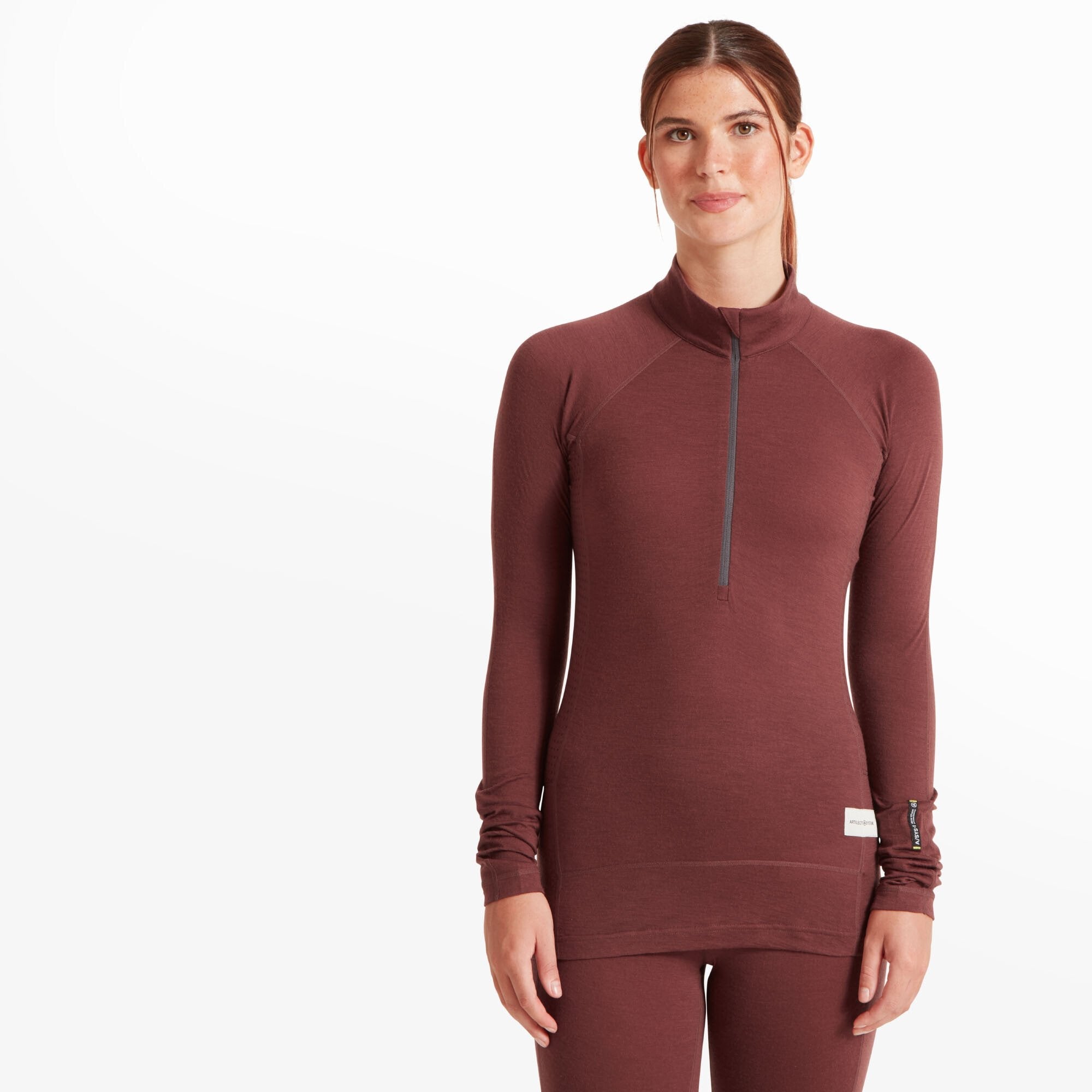 A woman with fair skin and brown hair tied back is modeling a Artilect Performance Womens Boulder 125 Quarter Zip Top Burgundy. The top has a snug fit, a high collar with a partial zipper, and thumbholes at the cuffs. The fabric appears to be soft and breathable. The model is standing against a plain white background, looking directly at the camera.