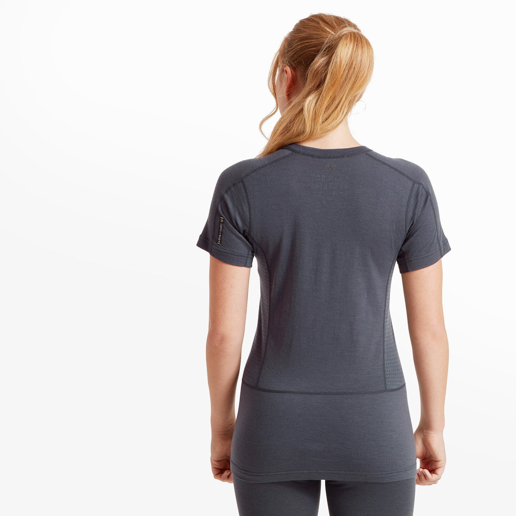 The woman is shown from the back, revealing the ergonomic paneling of the Artilect Performance Womens Boulder 125 Tee Blue, which has a slightly elongated back hem. A small brand logo is visible at the upper center, and the perforation details on the side panels continue along the back. Her blonde ponytail cascades down her back.