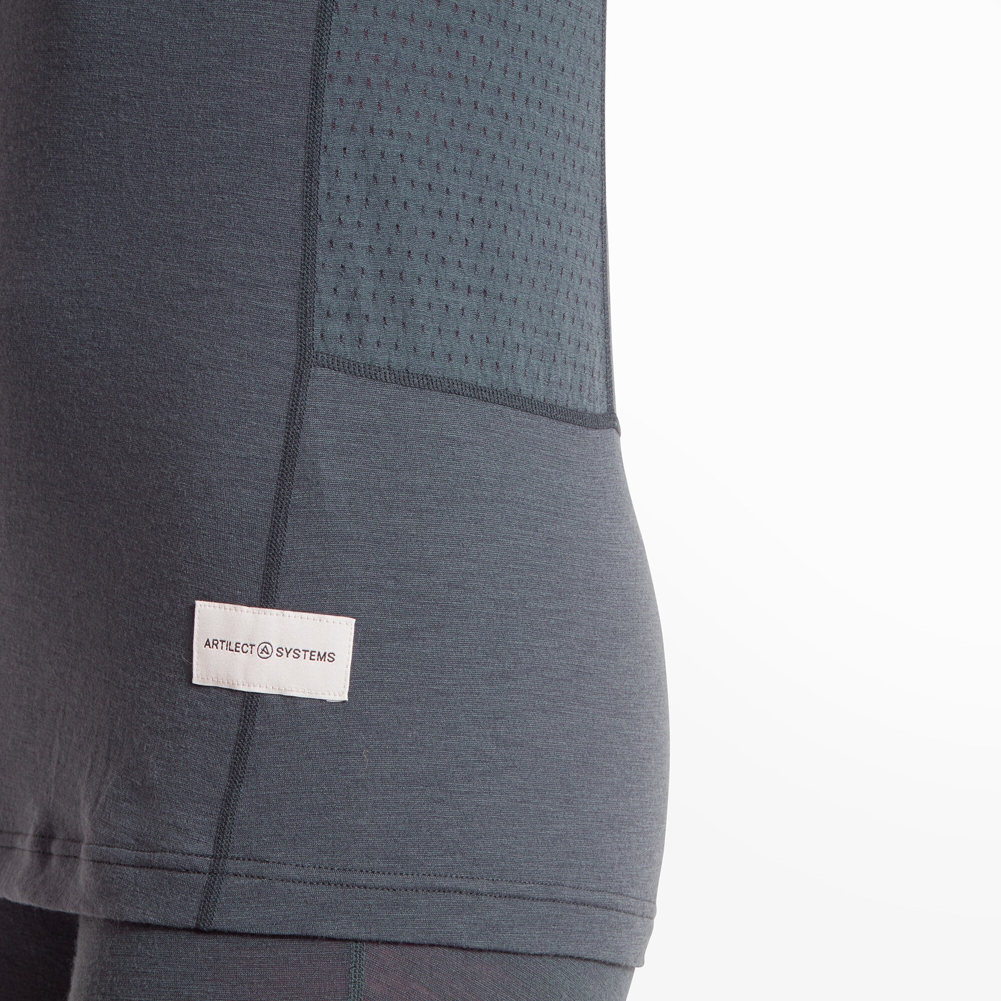 A detailed shot of the lower side of the Artilect Performance Womens Boulder 125 Tee Blue, focusing on the small white rectangular logo patch reading "ARTILECT SYSTEMS" and the perforated side panel for ventilation. The stitching and fabric texture are clearly visible.