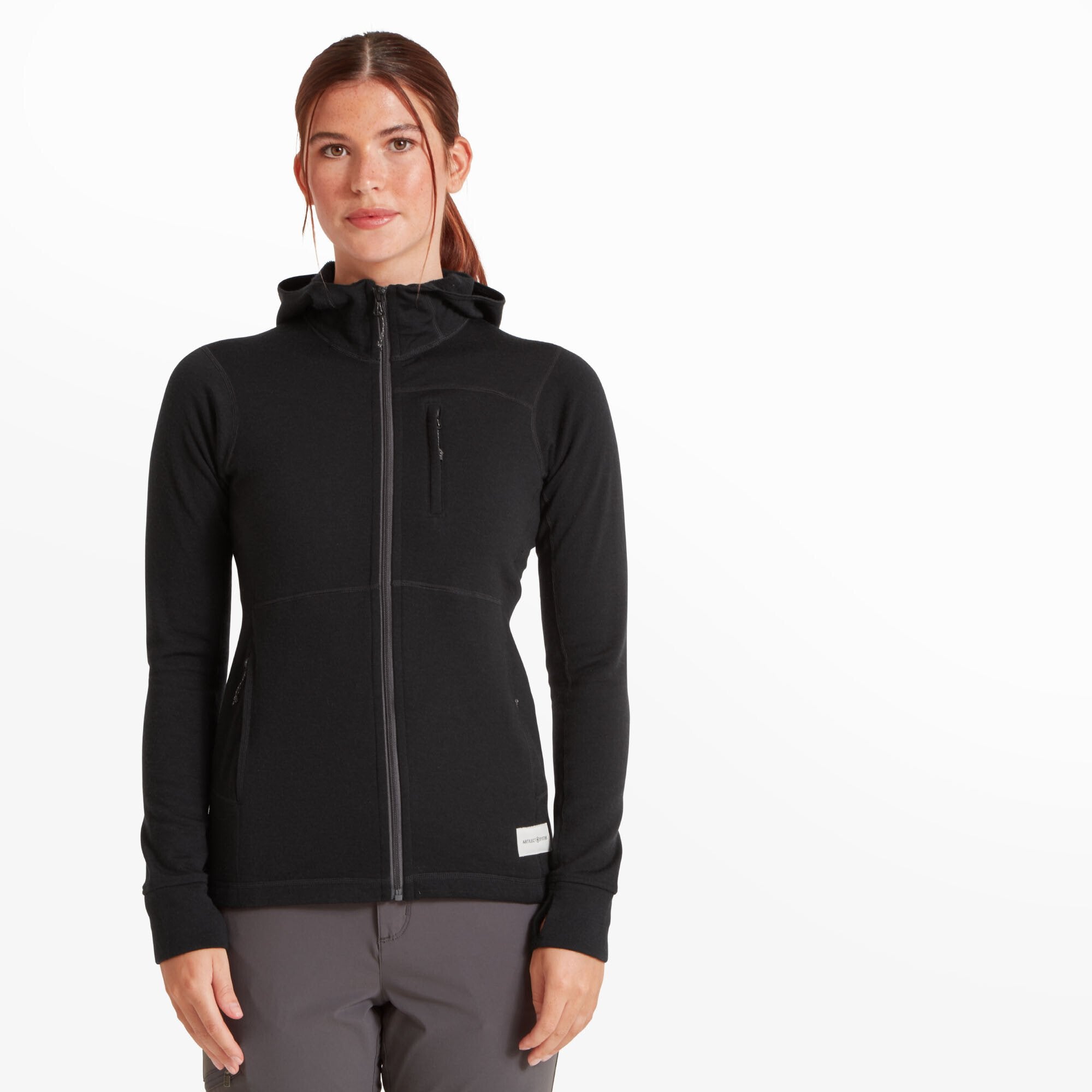 A woman is wearing a Artilect Performance Womens Eldorado Merino Hoodie Black made from merino wool. The hoodie features a high collar with an attached hood, a vertical zippered chest pocket, and two zippered hand pockets. The hem has a sewn-on white label that reads “ARTILECT SYSTEMS.” She is standing against a white background with her arms relaxed by her sides.