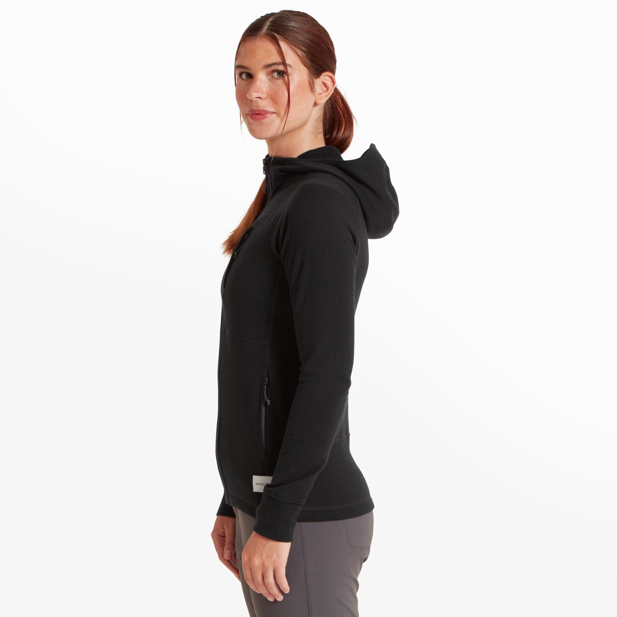 A profile shot of the same woman showing the left side of the Artilect Performance Womens Eldorado Merino Hoodie Black. The fitted silhouette highlights the athletic cut. The hood is shown resting flat on her back. The label and zipper details are partially visible on the left pocket, and the sleeves end with thumbhole cuffs.