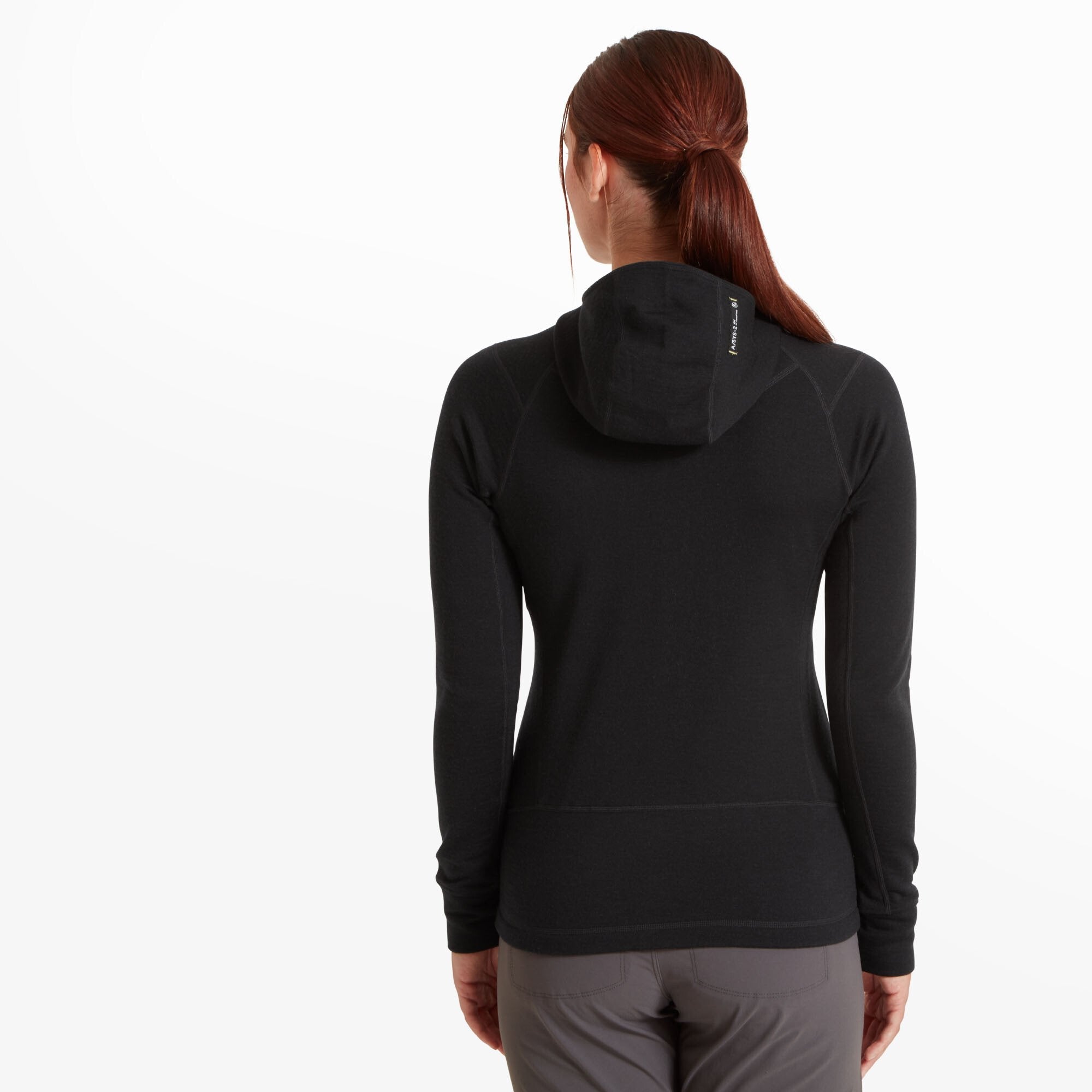 The woman is facing away from the camera, revealing the back design of the Artilect Performance Womens Eldorado Merino Hoodie Black. The hood is up, lying flat against the back, and subtle seam lines contour the upper back. The fit hugs the body, and the sleeve and bottom hem lines are clean and minimal.