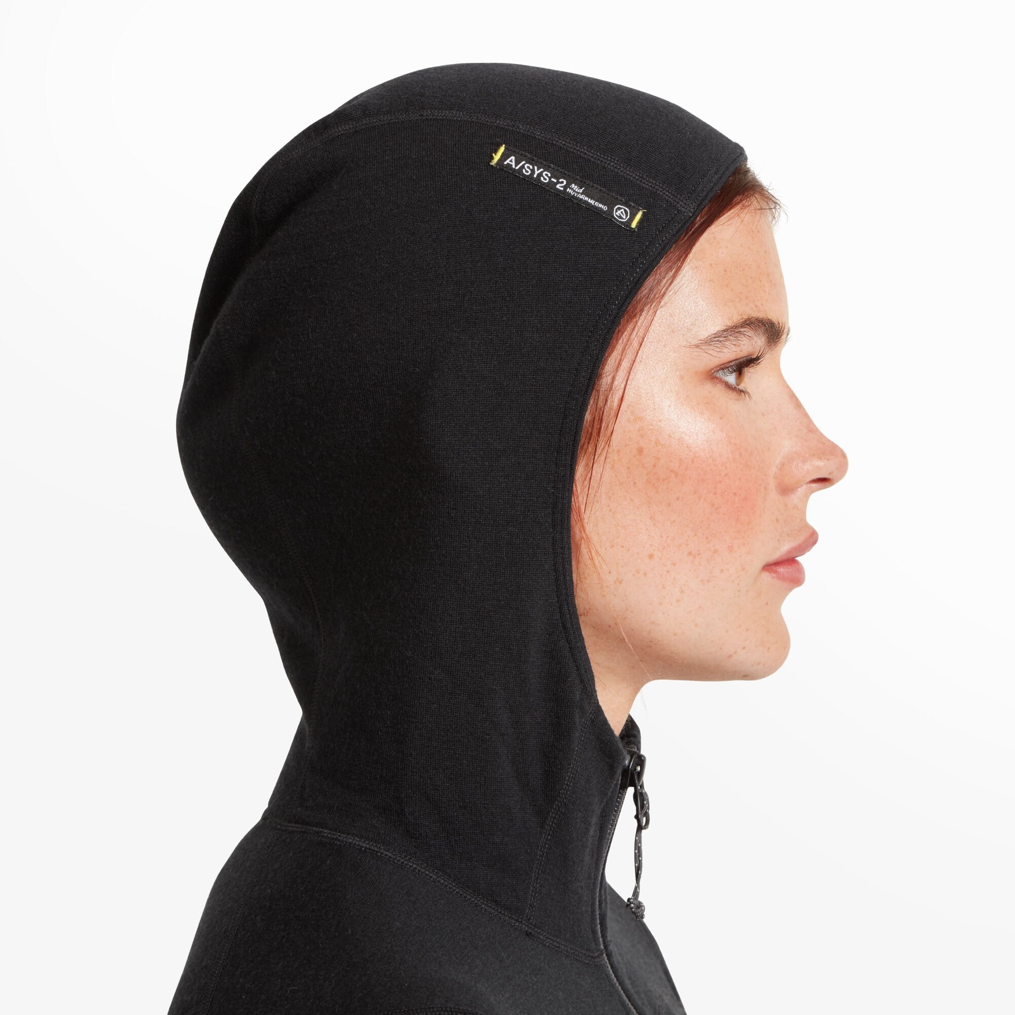 A detailed side profile close-up of the model’s face and hood on the Artilect Performance Womens Eldorado Merino Hoodie Black. The hood fits snugly and features a small printed label reading “A/SYS-2 by Artilect” with a merino wool insignia. The material appears soft and stretchy, suitable for layering.