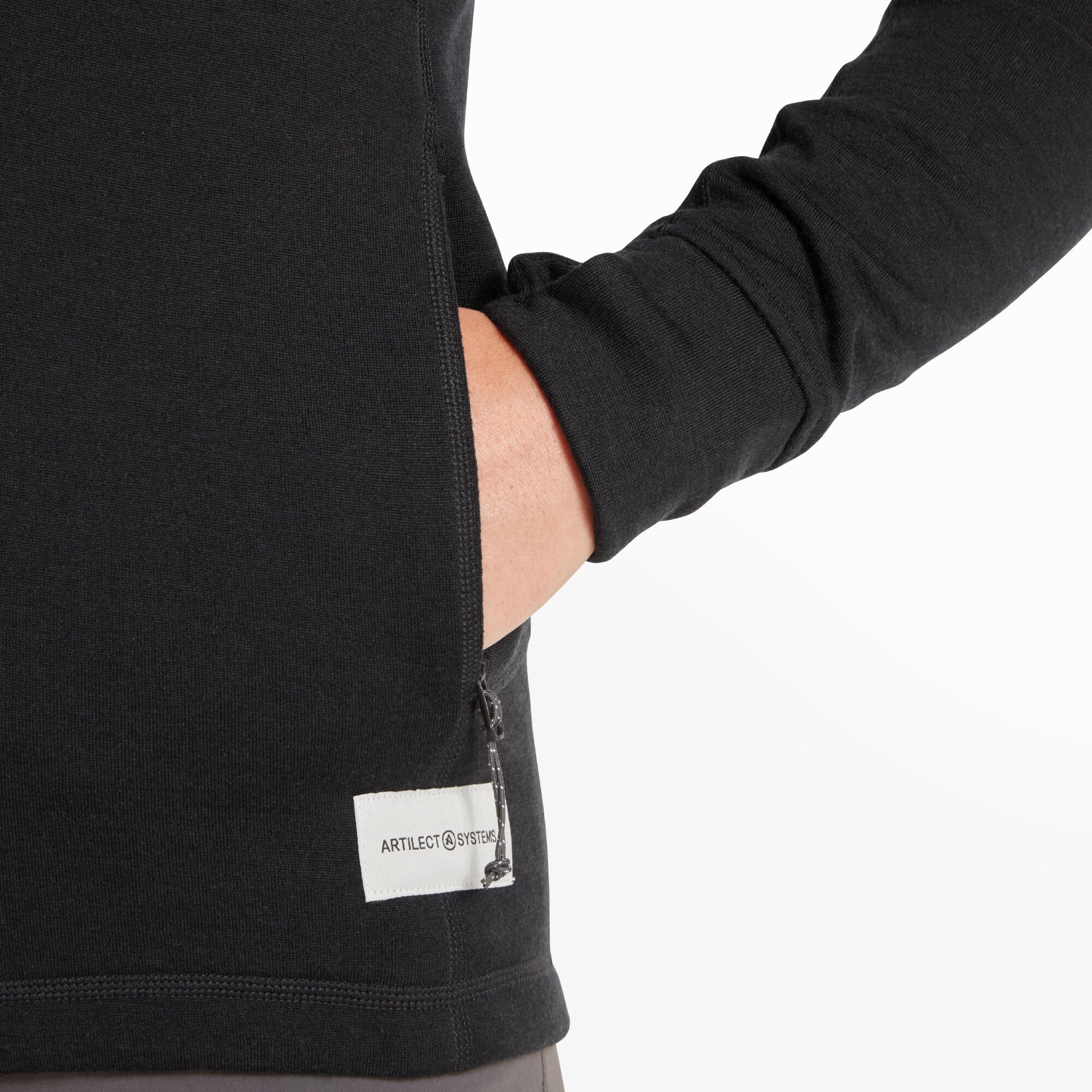 Detailed shot of the model’s hand in the zippered left pocket on the Artilect Performance Womens Eldorado Merino Hoodie Black. The branded white ARTILECT SYSTEMS tag is sewn into the hem nearby, and the knit texture of the fabric is clearly visible.