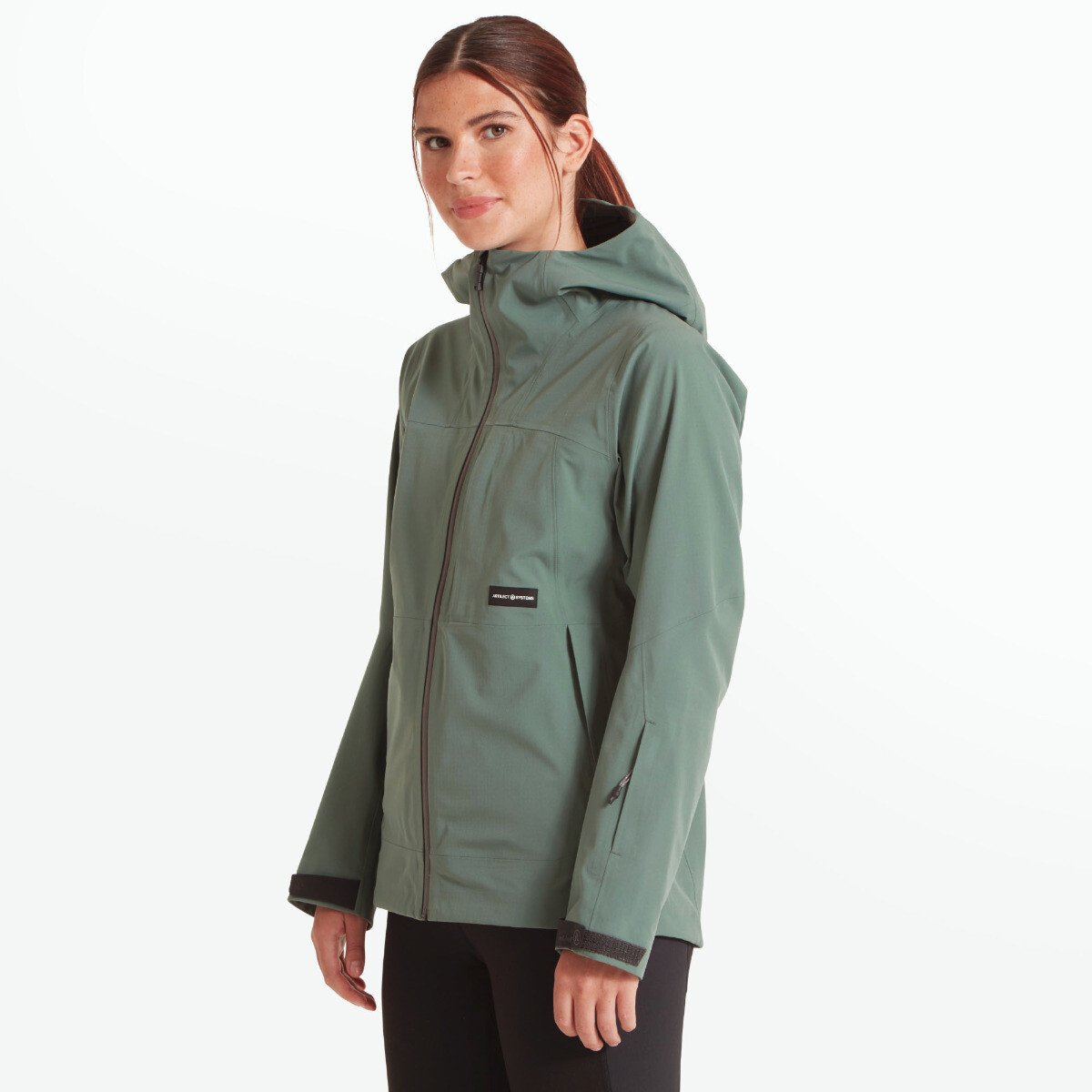 The model is turned slightly to her left, offering a clear side profile of the Artilect Performance Womens Grandview 3L Jacket Green’s tailored fit. The articulated sleeves and paneling around the torso are visible, emphasizing mobility. The hood is neatly shaped and fits close to the head.