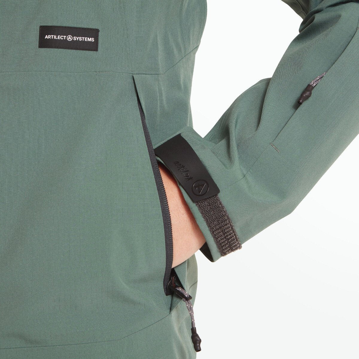 The model’s hand is partially inside the side zippered pocket. Velcro cuff tabs with rubberized Artilect branding are visible. The fabric texture and detail of the seams around the midsection are clearly shown on the Artilect Performance Womens Grandview 3L Jacket Green.