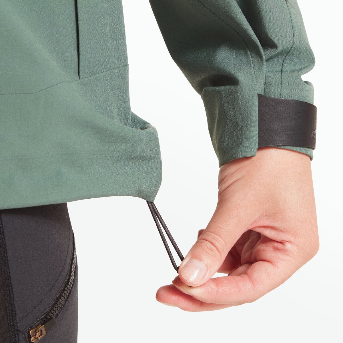 A detail image showing the model’s hand pulling on the dual cord lock at the hem, demonstrating how the bottom of the Artilect Performance Womens Grandview 3L Jacket Green can be cinched to block wind or snow.