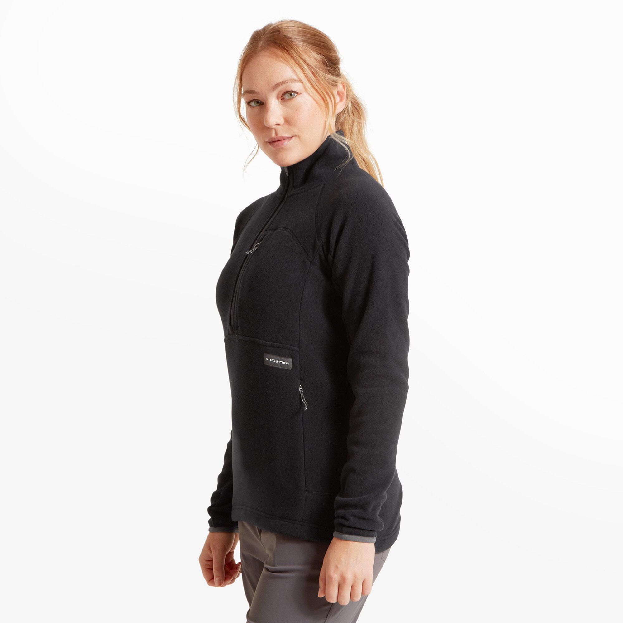 A woman wearing a Artilect Performance Womens Halfmoon Bio Pullover Black stands facing forward with a soft smile. The fleece top features a half-zip design with a chest pocket zipper and two hand pockets. She wears it over gray pants and has her blonde hair tied back.