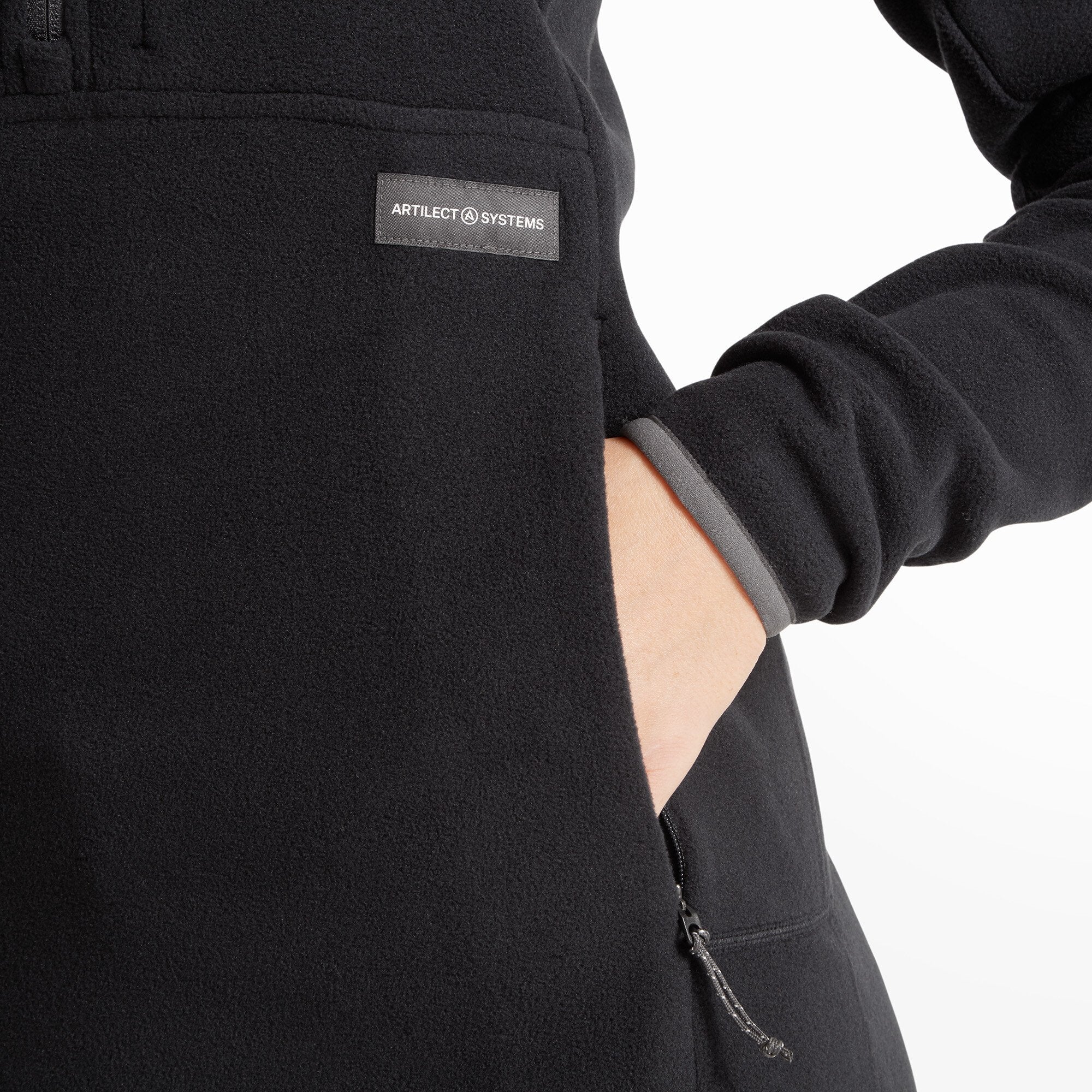 Zoomed-in view of the left pocket area of the Artilect Performance Womens Halfmoon Bio Pullover Black. A hand is partially inserted into the fleece-lined pocket. A black “ARTILECT SYSTEMS” label is sewn just above the zipper.