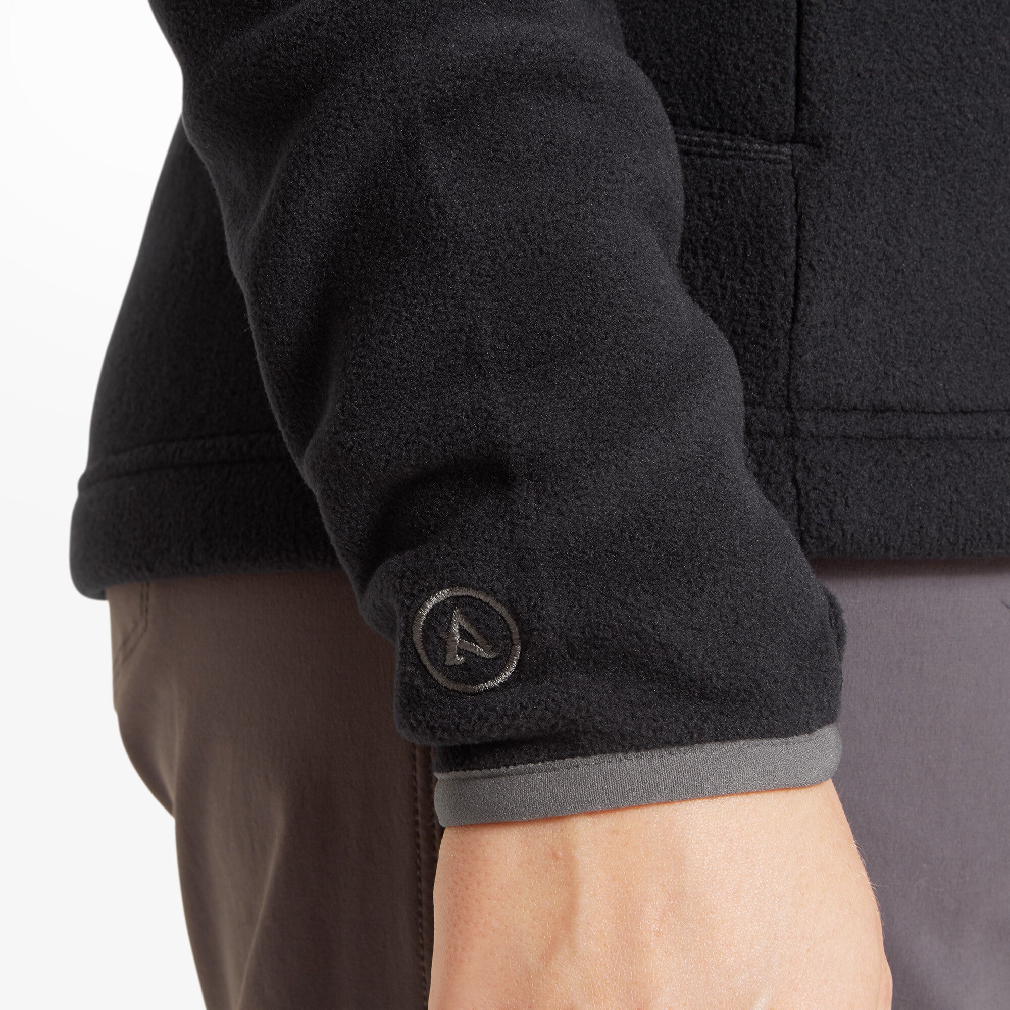 Close-up of the sleeve cuff of the Artilect Performance Womens Halfmoon Bio Pullover Black. The woman’s arm is relaxed at her side, displaying the gray trim at the wrist and an embroidered Artilect “A” logo in light gray near the cuff.