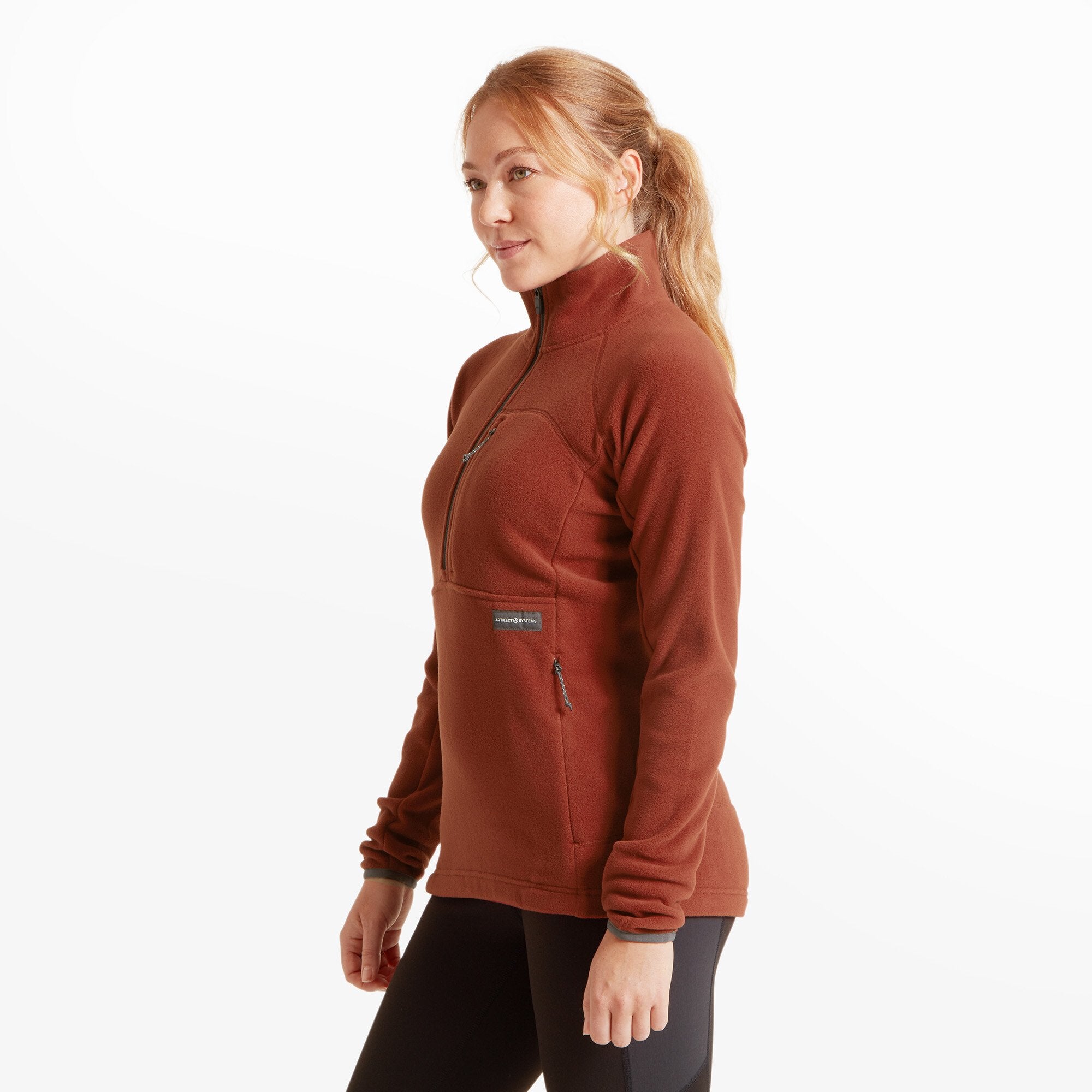 The model is turned slightly to her left, giving a three-quarter view that highlights the Artilect Performance Womens Halfmoon Bio Pullover Orange’s fitted silhouette and zippered side pocket on the right. The soft fleece texture and structured cut are clearly visible, emphasizing both comfort and performance style.