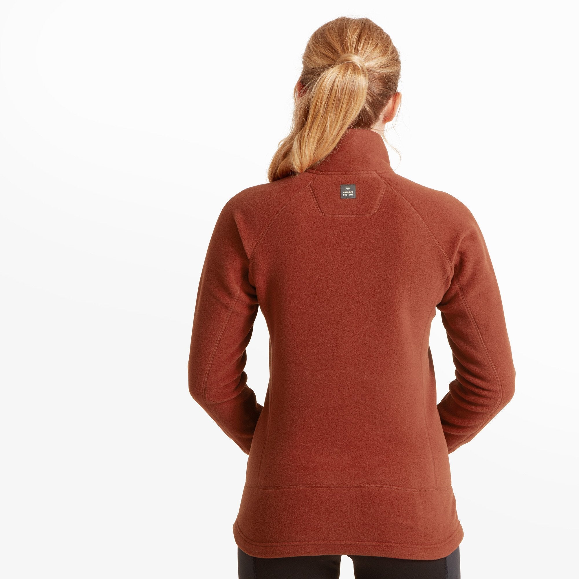 A rear view of the model showcases the back design of the Artilect Performance Womens Halfmoon Bio Pullover Orange, which includes a high collar and simple seam construction that tapers at the waist. A small rectangular Artilect logo patch is centered just below the collar.