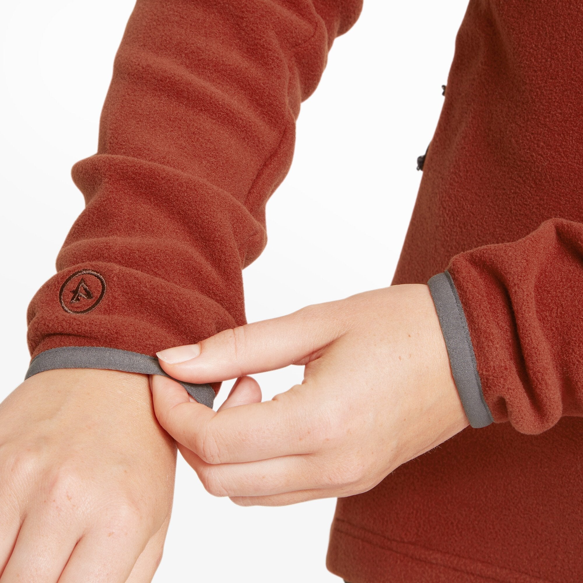 Close-up of both of the model’s hands adjusting the cuff of the Artilect Performance Womens Halfmoon Bio Pullover Orange. The inner grey lining is revealed as the cuff is rolled back, and a small, circular embroidered Artilect logo is visible on the left sleeve near the wrist.